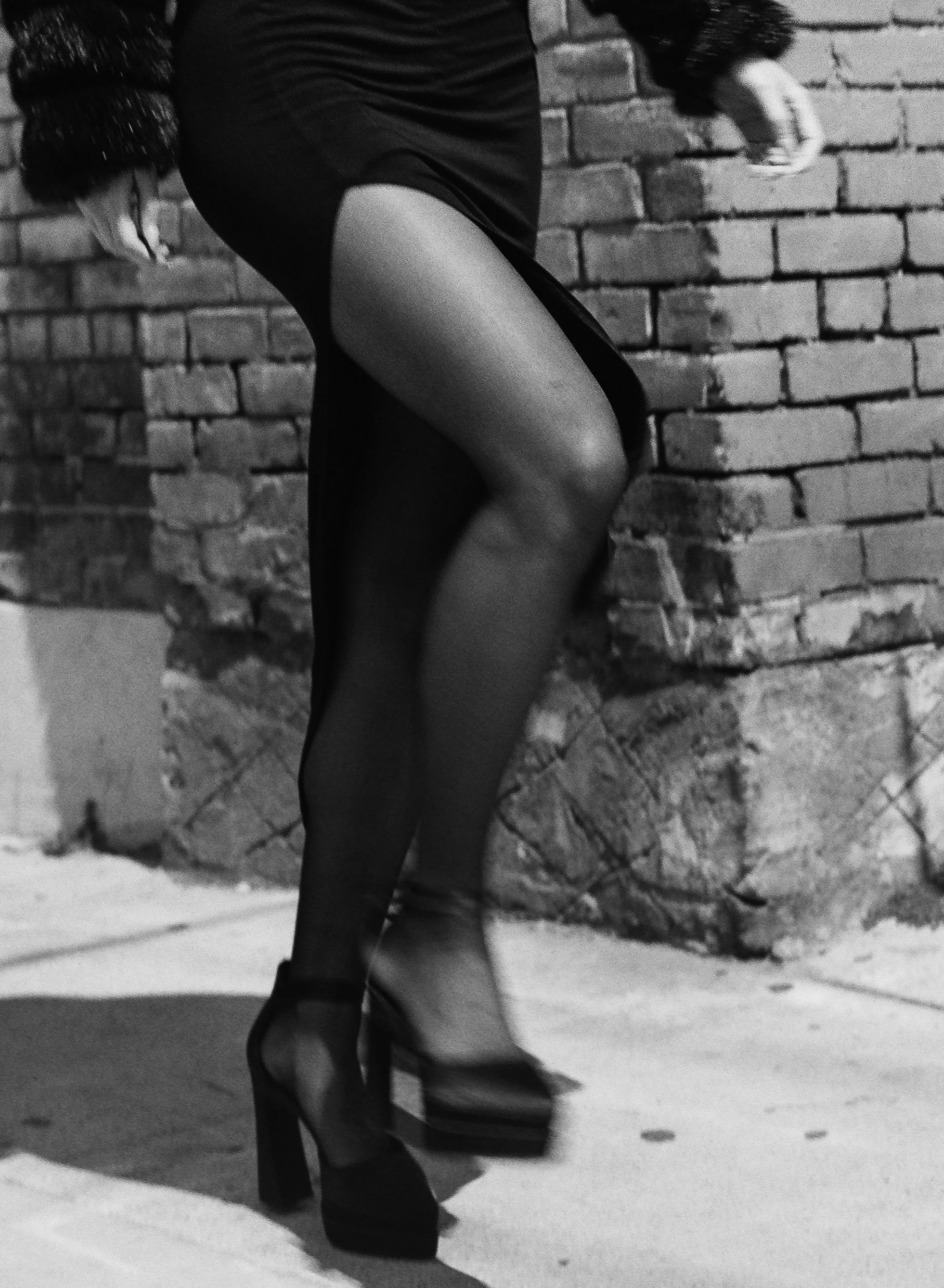 A woman wearing high heels, a sheer black dress, and gloves, standing against a brick wall. The image is in black and white.
