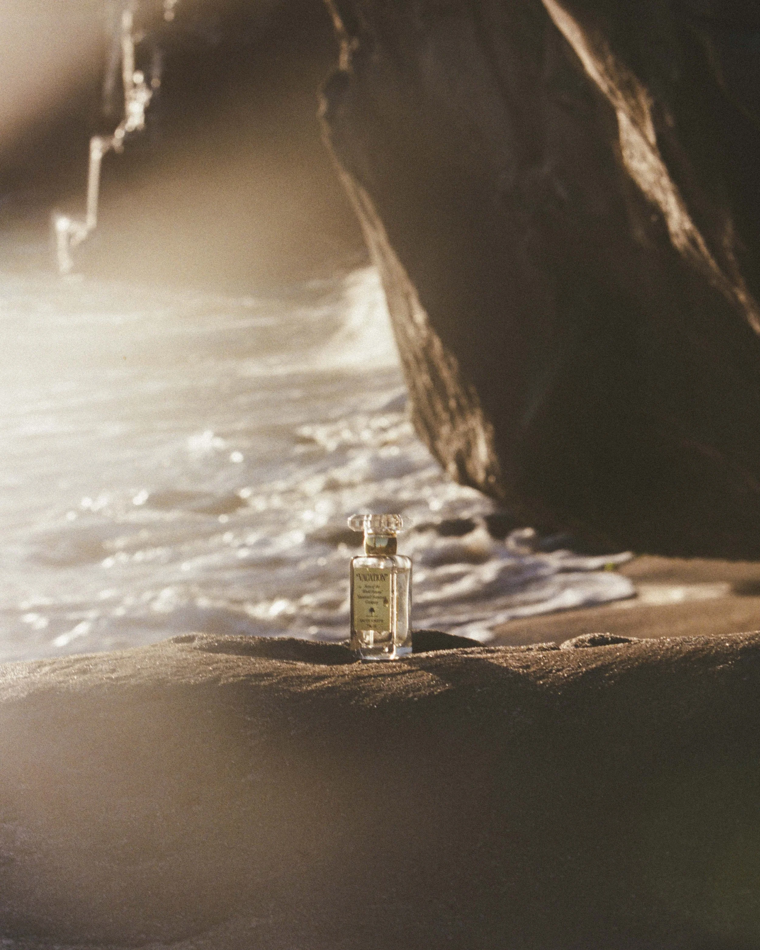 A small glass bottle of perfume placed on the sand at a beach, with a large rock formation and the ocean in the background.