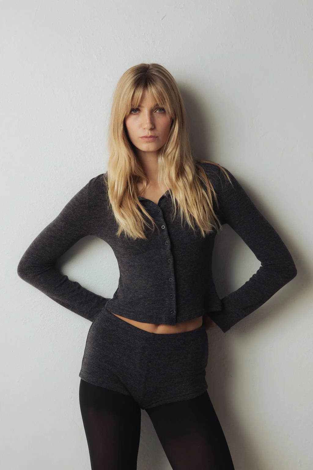 Modeling portfolio image of a girl against a white wall, wearing a dark gray cardigan, matching shorts, and black leggings.