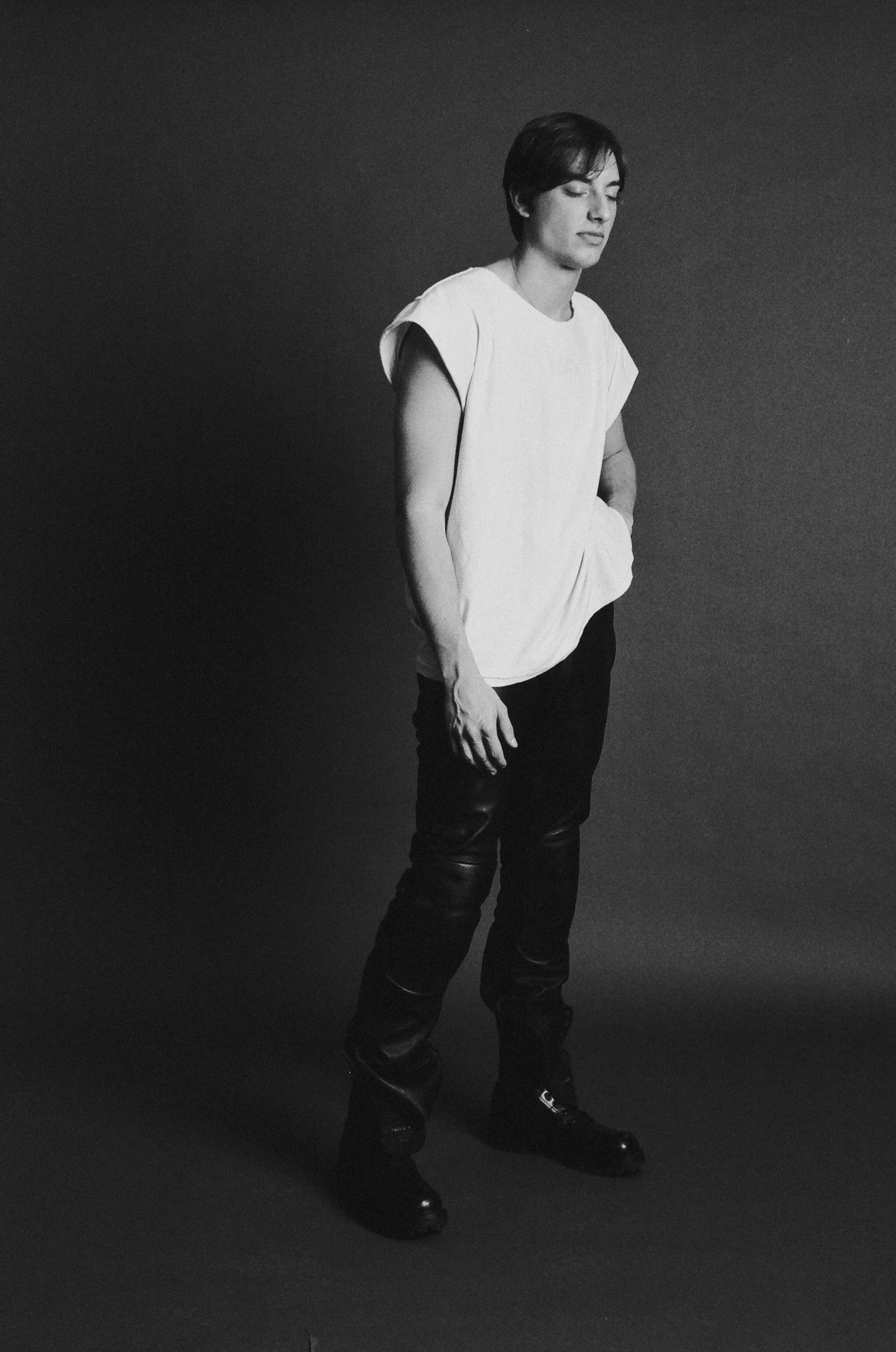 Black and white photo of a person with short hair, wearing a sleeveless white top, black pants, and black boots, standing against a plain dark background.