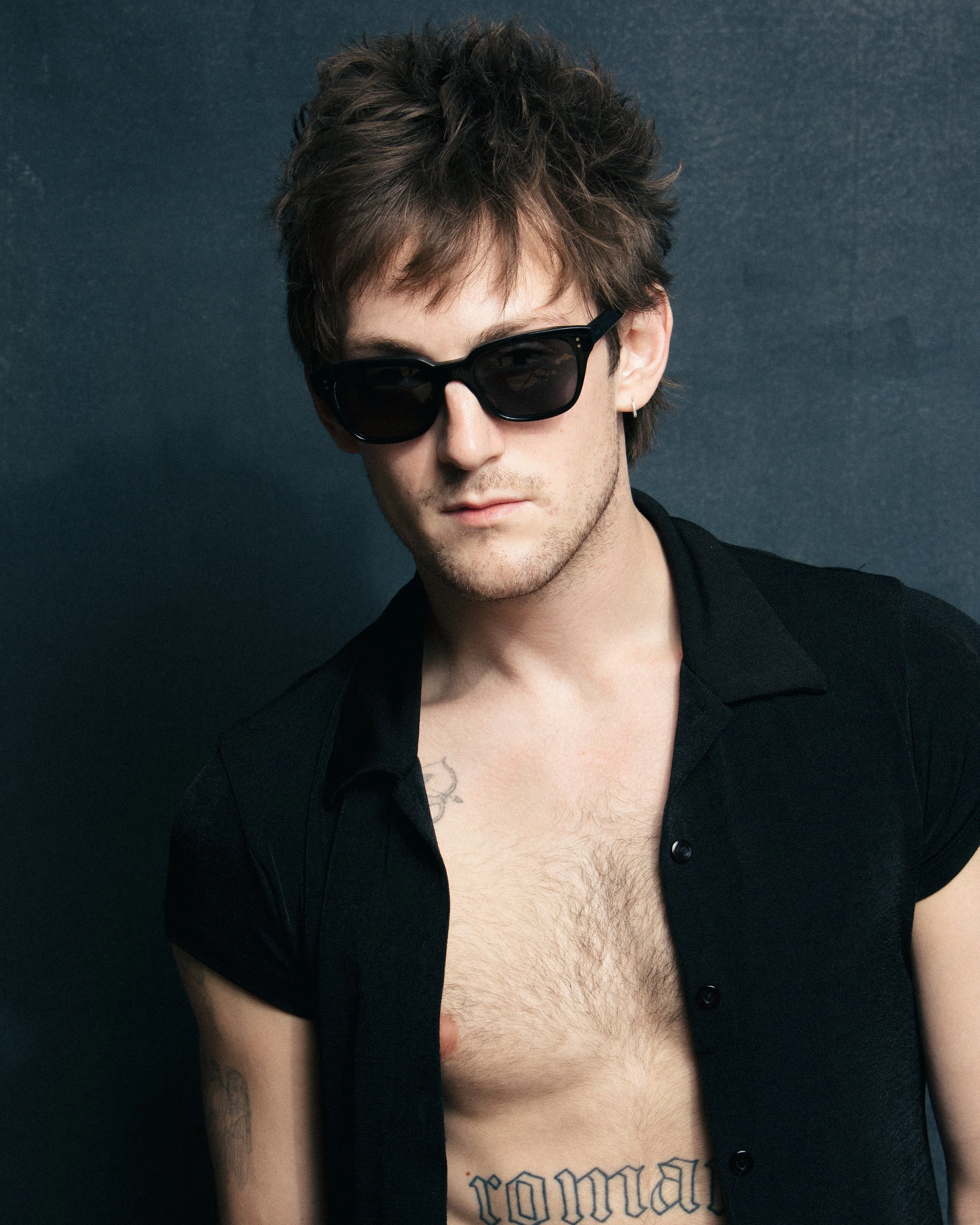 Young man with tousled brown hair wearing black sunglasses, a black short-sleeve button-up shirt, and a tattoo that reads 'romantic' on his chest, against a dark background.
