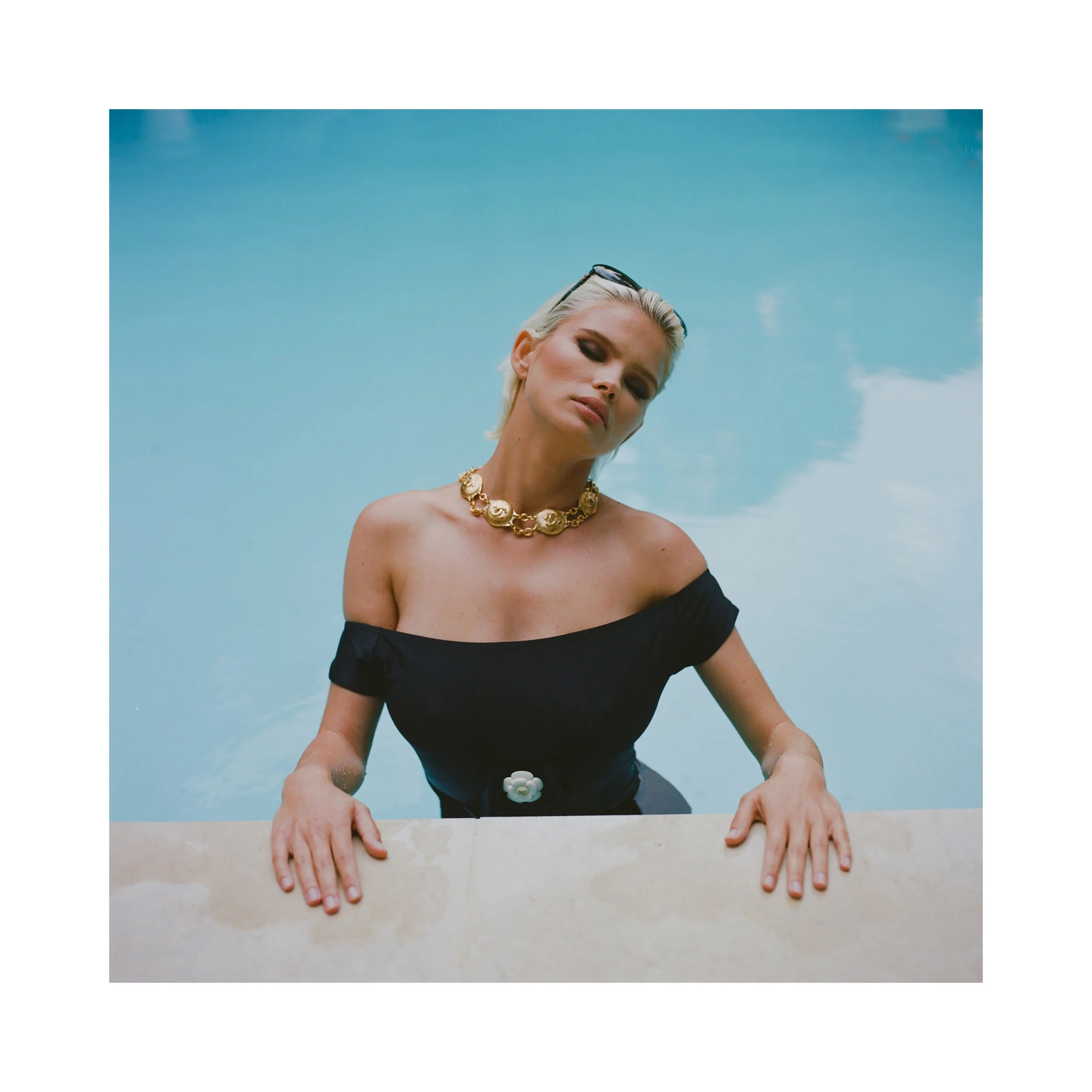 Woman with blonde hair wearing a black off-shoulder top and gold necklace, relaxing in a swimming pool with a blue sky reflected in the water.