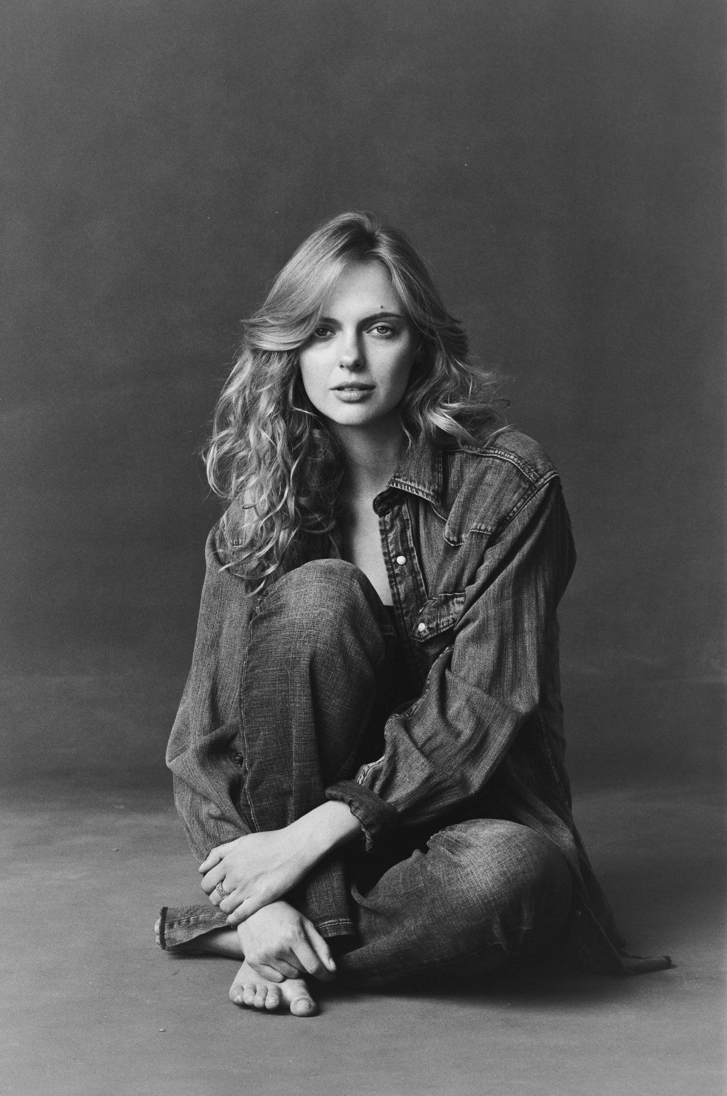 Black and white photo of a woman sitting on the floor with her legs crossed, wearing a denim jacket and jeans, with long wavy hair and a neutral expression. Actress, Jessica Belkin, Acting Headshots