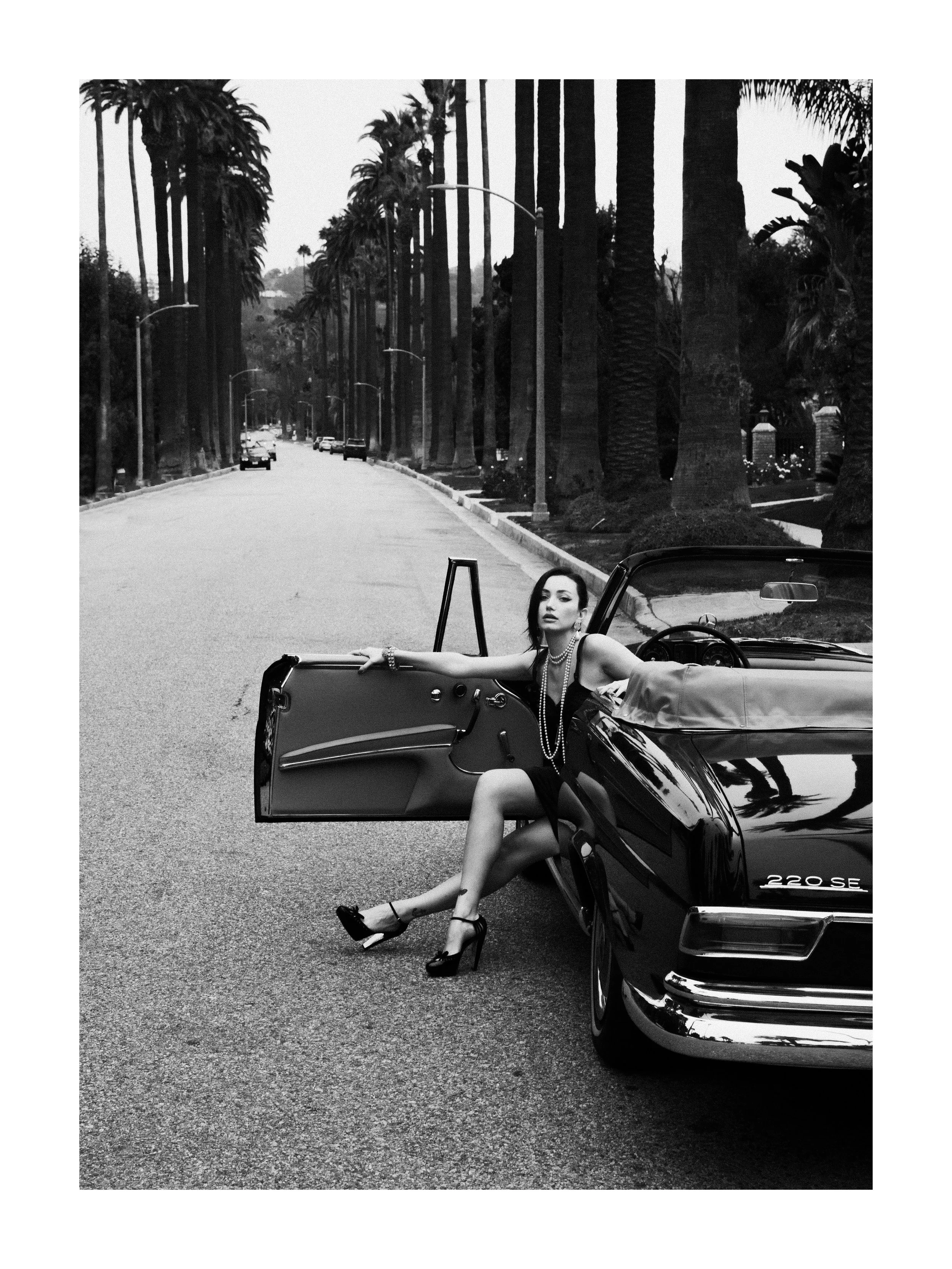 A woman in a glamorous dress and high heels sitting in a vintage convertible car, parked on a palm tree-lined street, black and white photo.