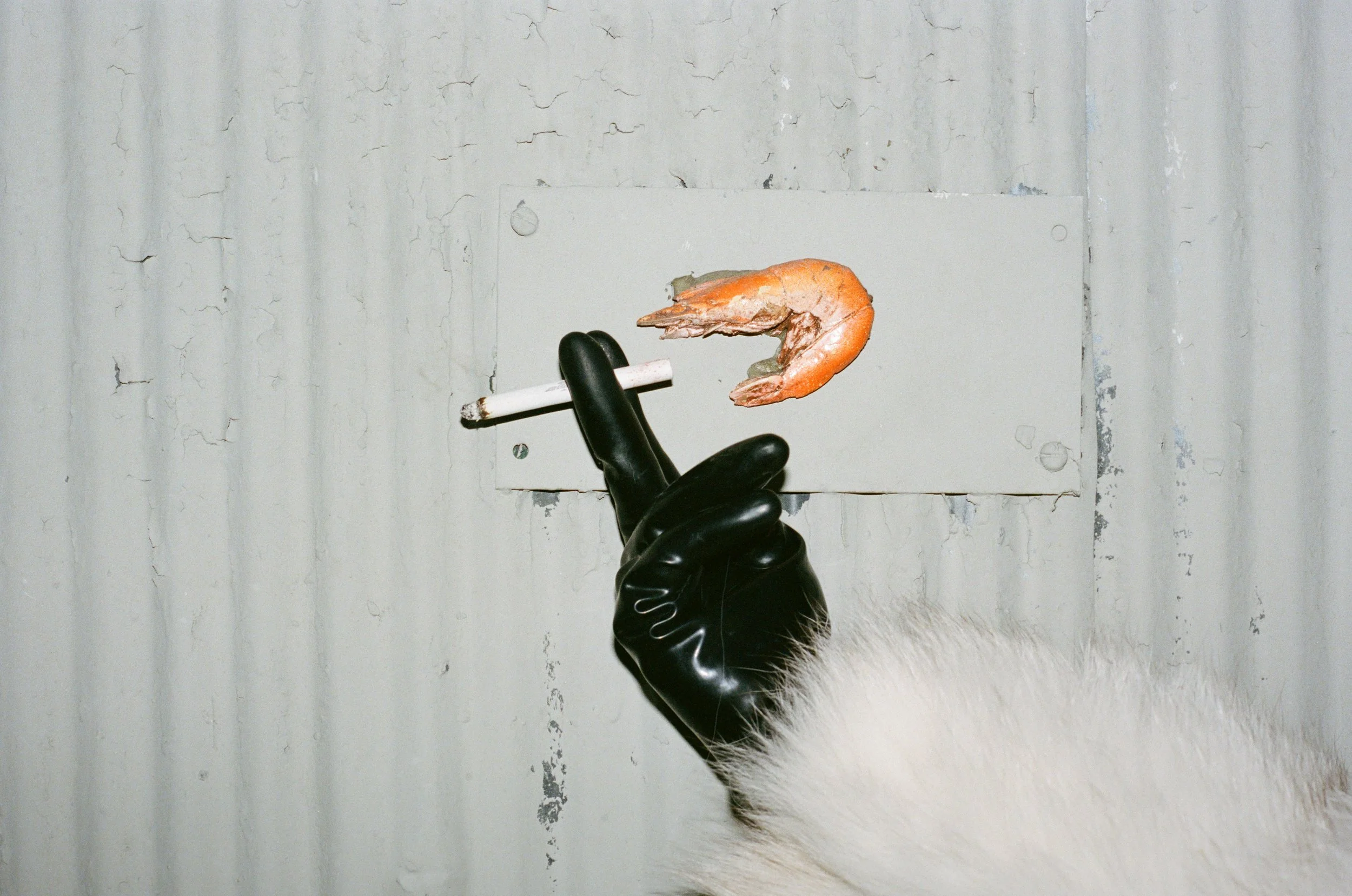 A gloved hand with white fur on the wrist holding a cigarette with a shrimp on it in front of a corroded metal wall.