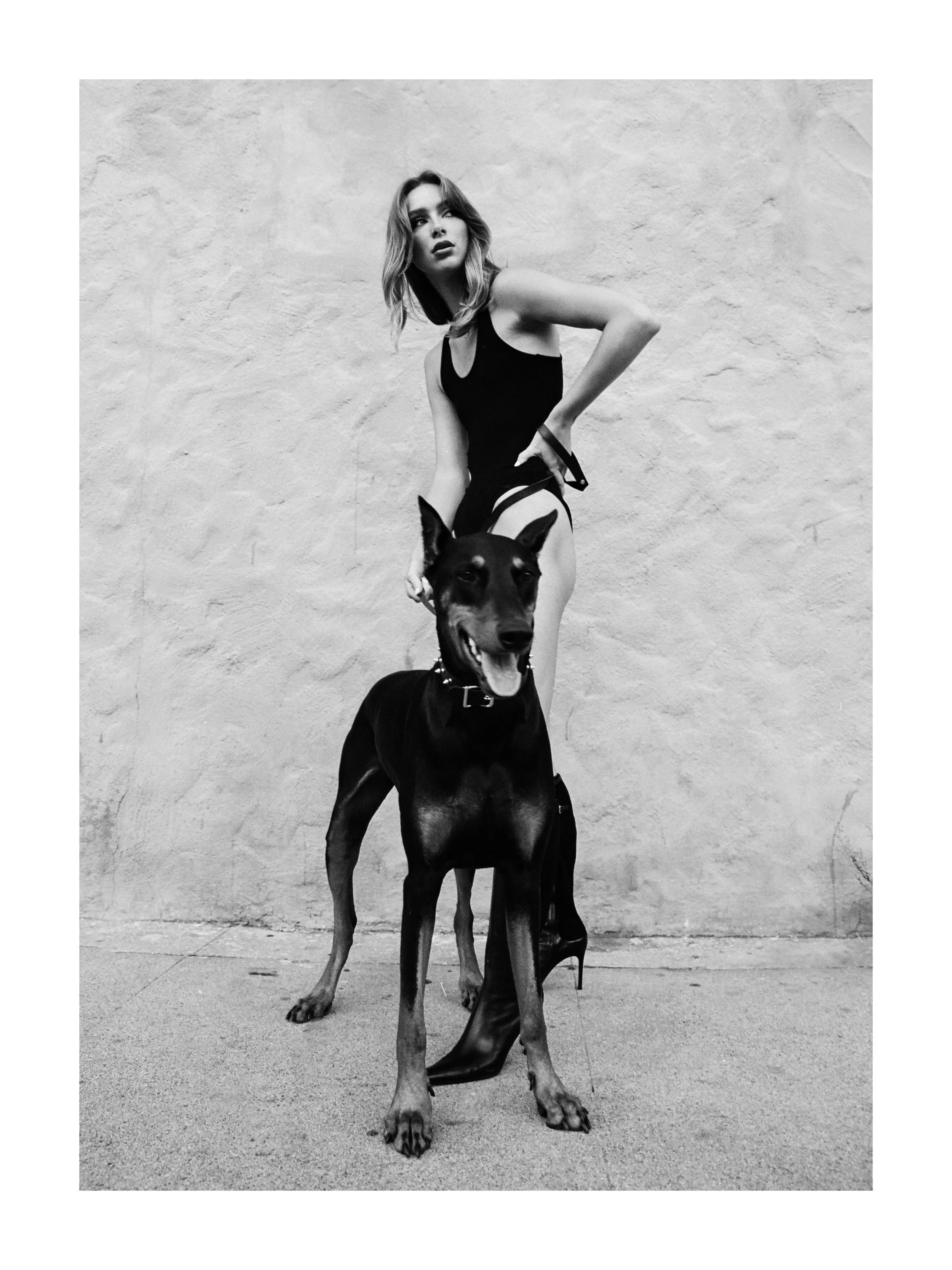 A woman in a black outfit with high heels and a dog with a harness standing against a plain wall.