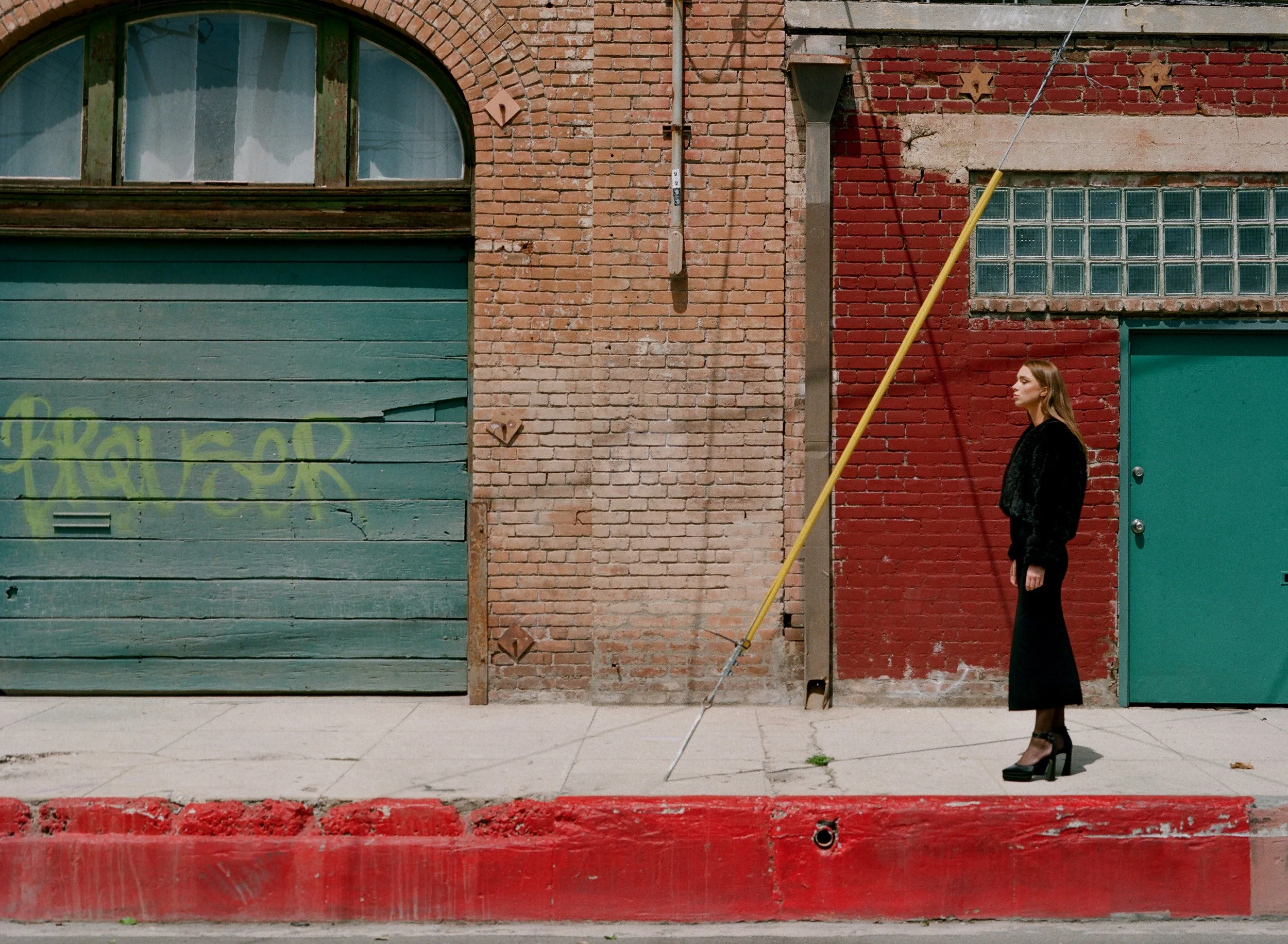 A young woman in black clothing standing on a city sidewalk in front of a colorful brick building with a large green garage door and a smaller green door, holding a yellow painter's pole.