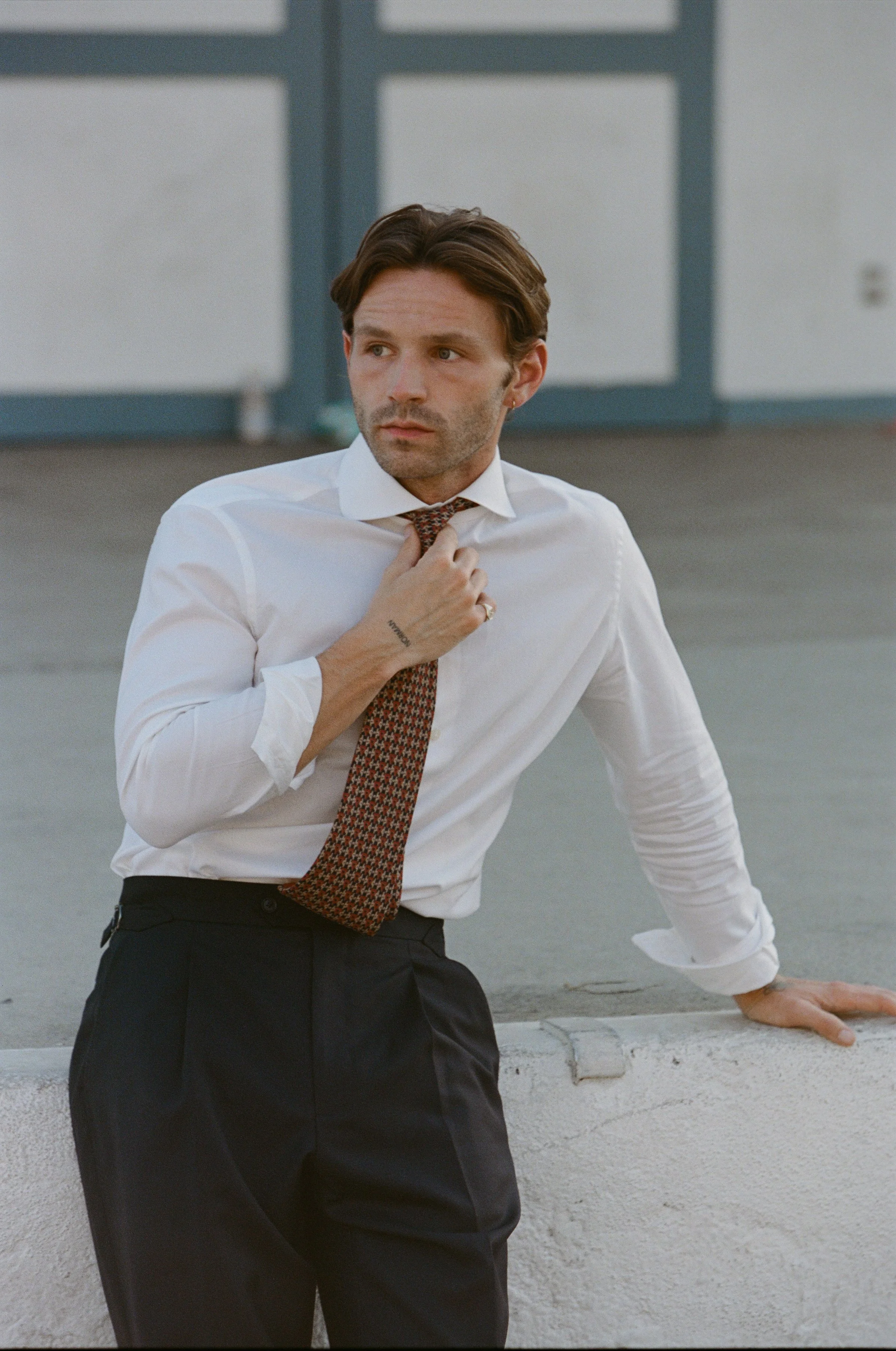 A man wearing a white dress shirt and patterned tie, leaning against a concrete barrier, adjusting his necktie, in an outdoor urban setting.