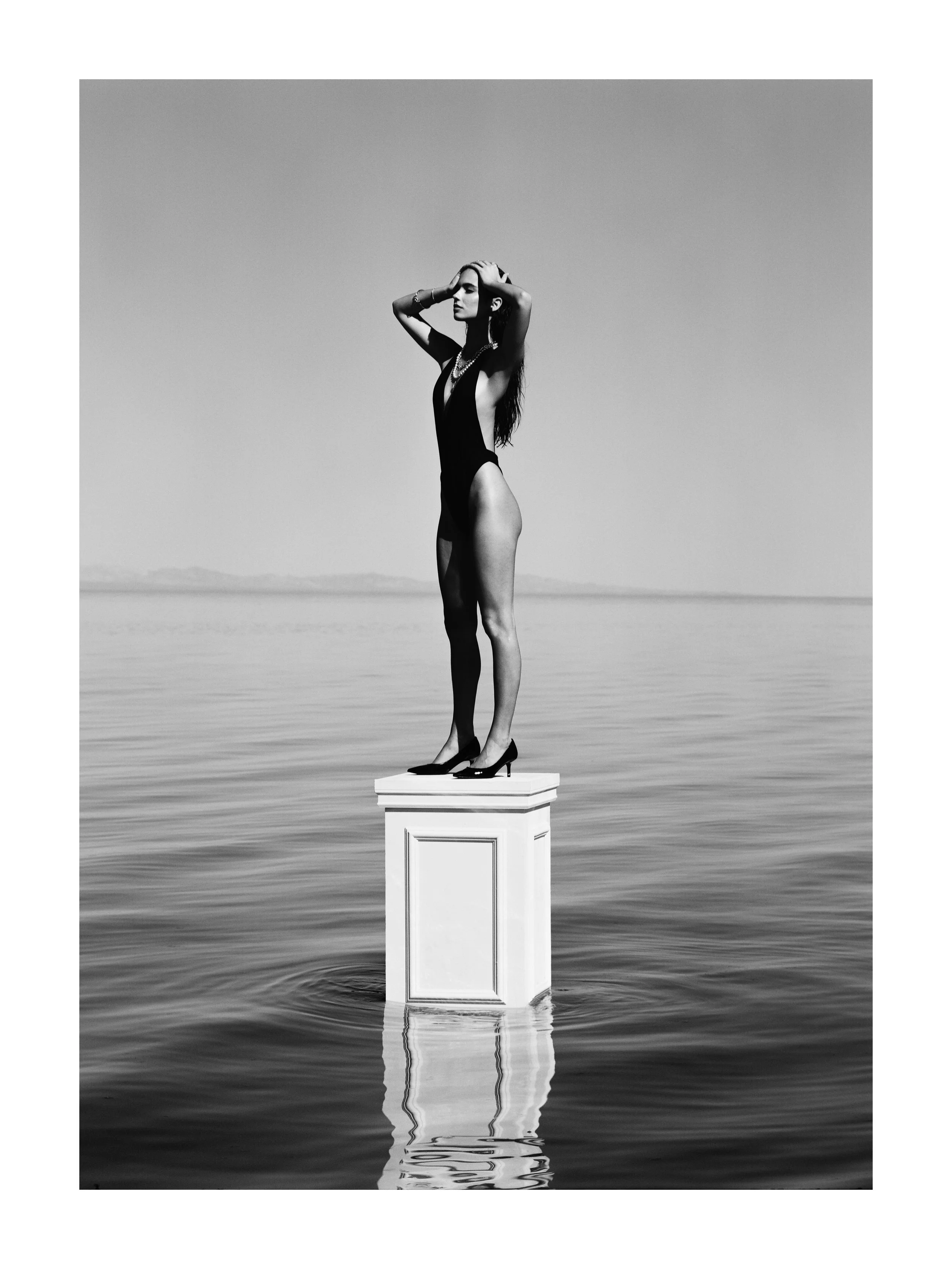 A woman in a swimsuit standing on a pedestal in the water with a vast, calm ocean and distant mountains in the background, captured in black and white.