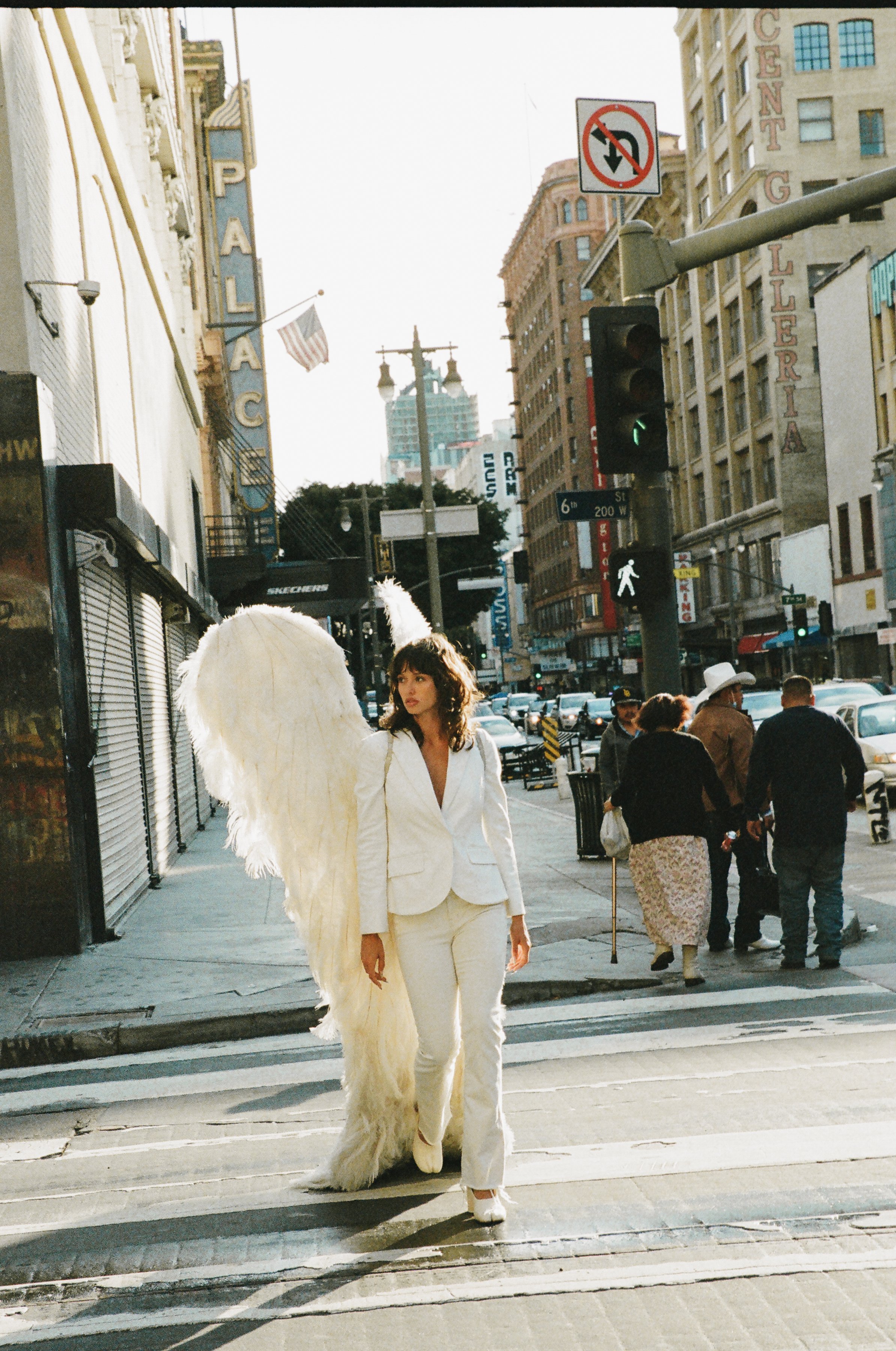 Woman dressed as an angel with white wings and a white suit walking across a city crosswalk, with pedestrians and buildings visible.