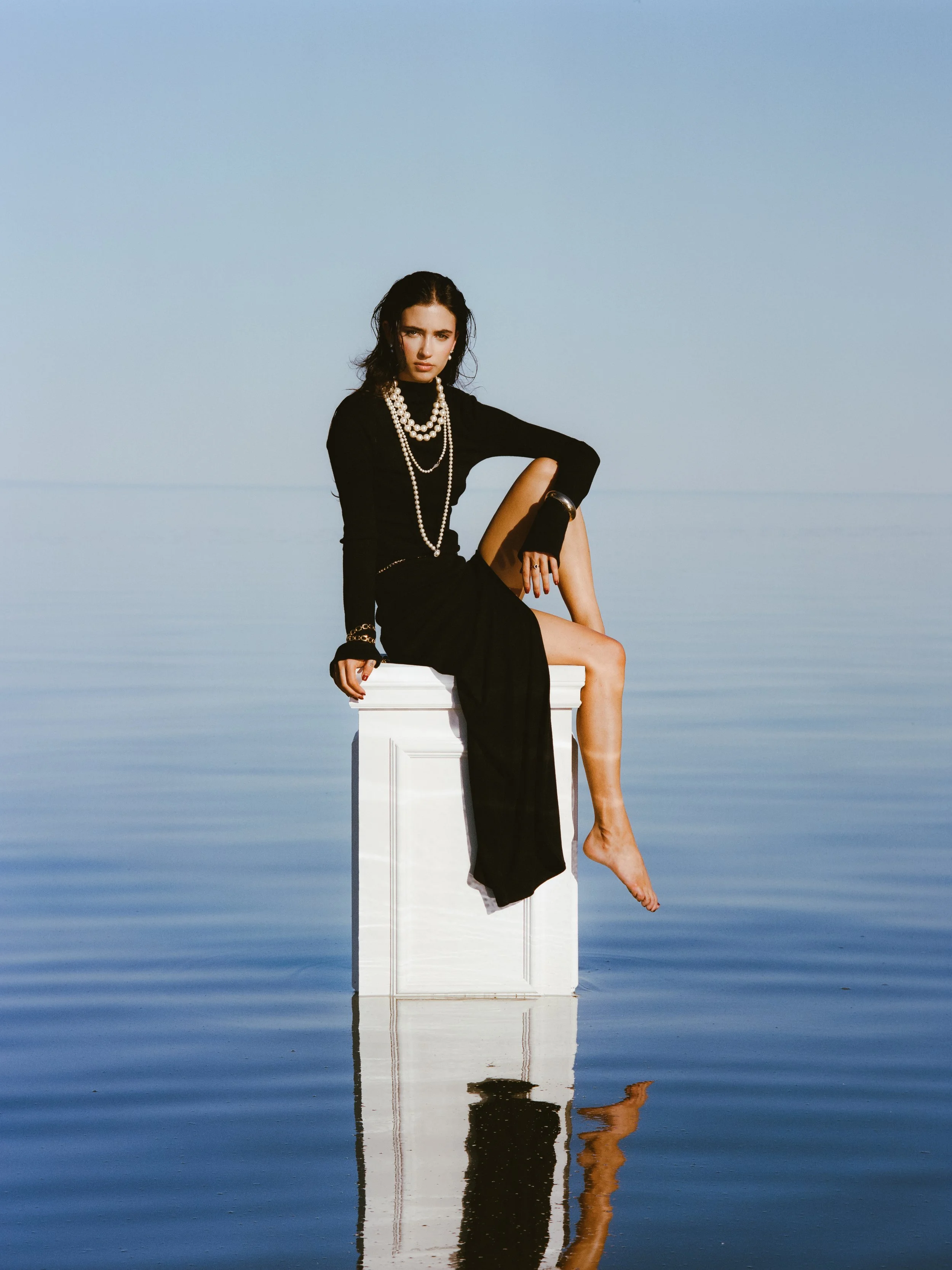 A woman in a black dress with jewelry sitting on a white pedestal in water with a blue sky background.