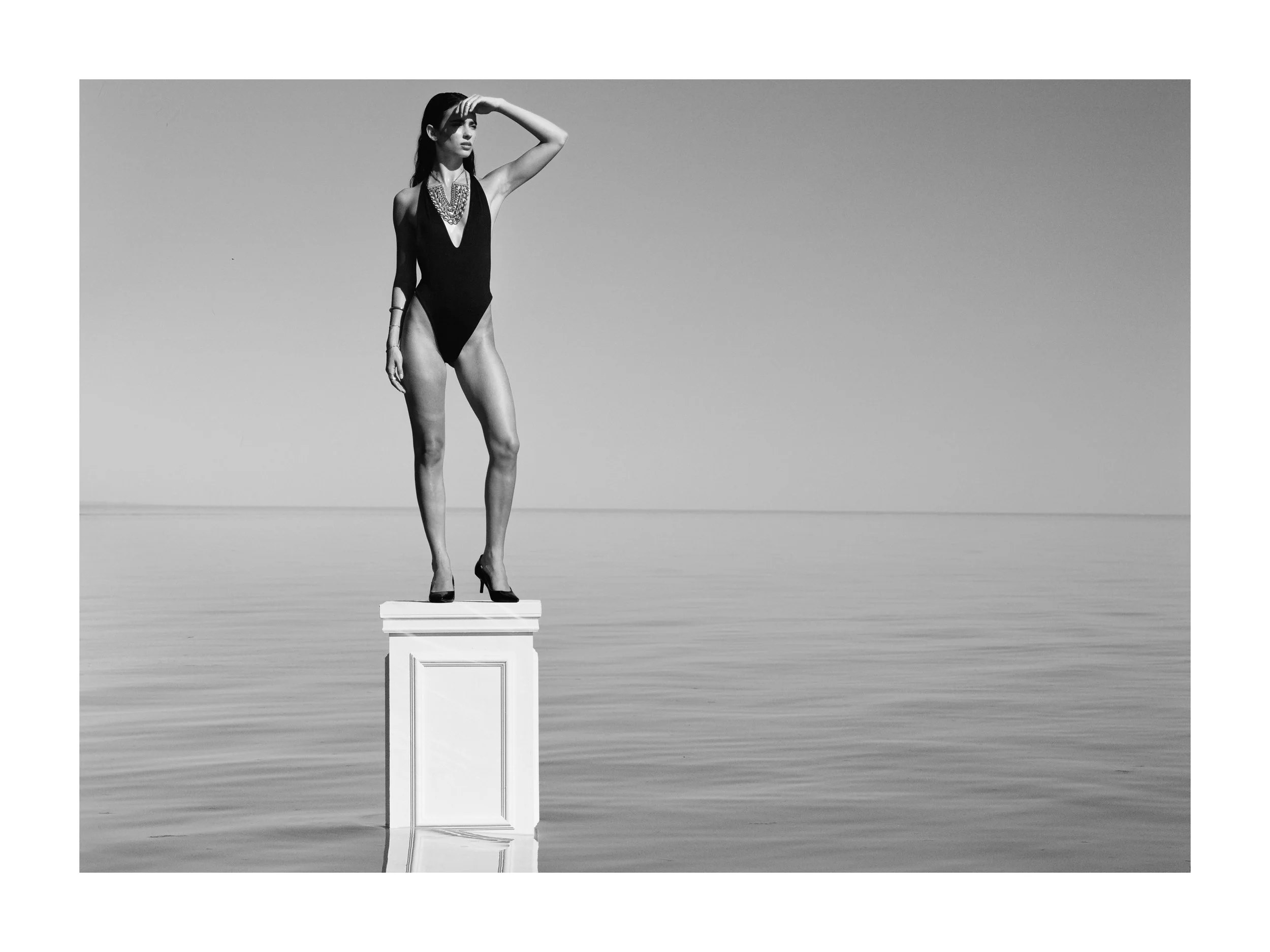 A woman in a black swimsuit and heels stands on a pedestal in water, looking into the distance with her hand shading her eyes, black and white photo.