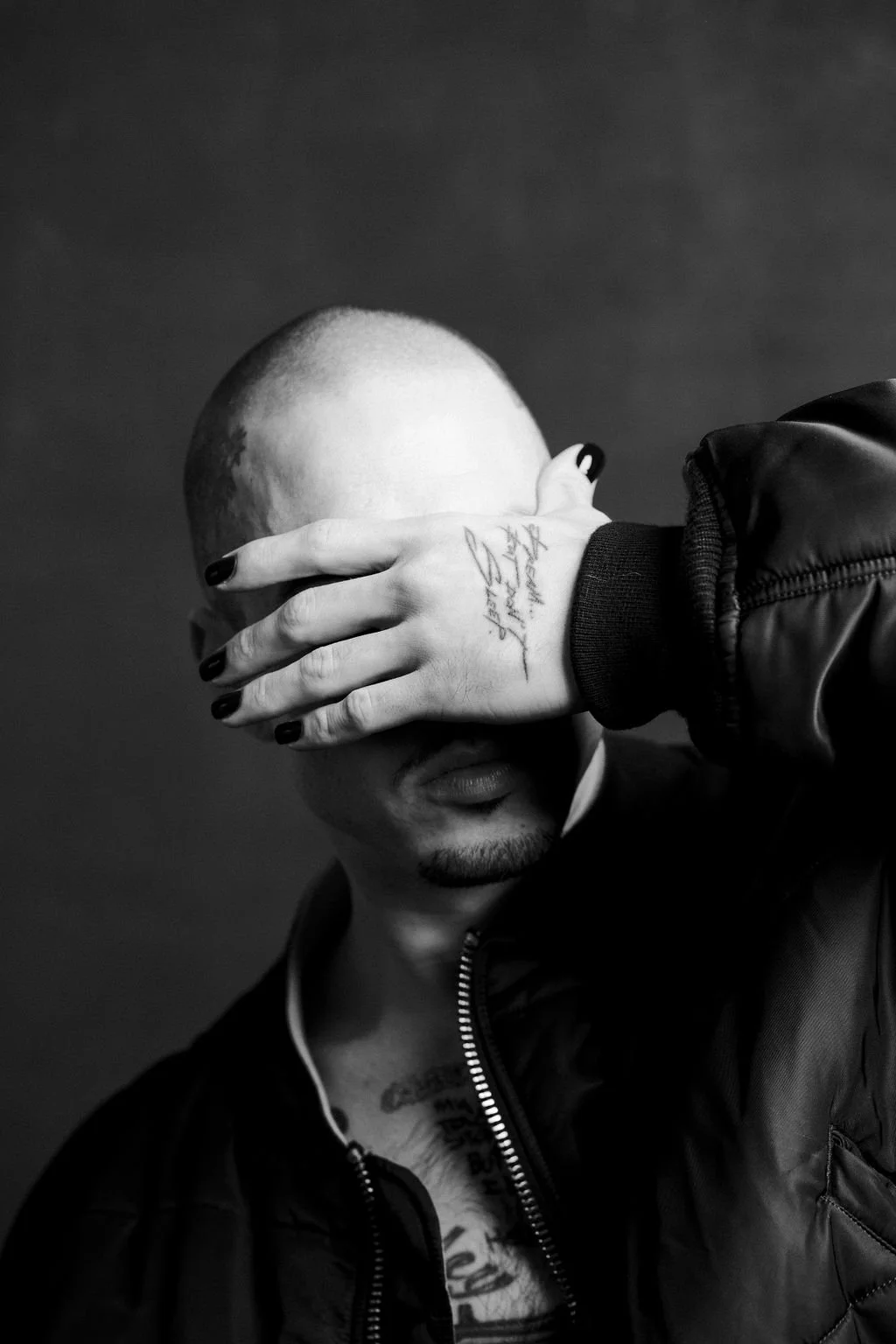 A black and white photo of Mitch Modes covering his eyes with his left hand that has tattoos and black nail polish, wearing a jacket with a zipper and a necklace.