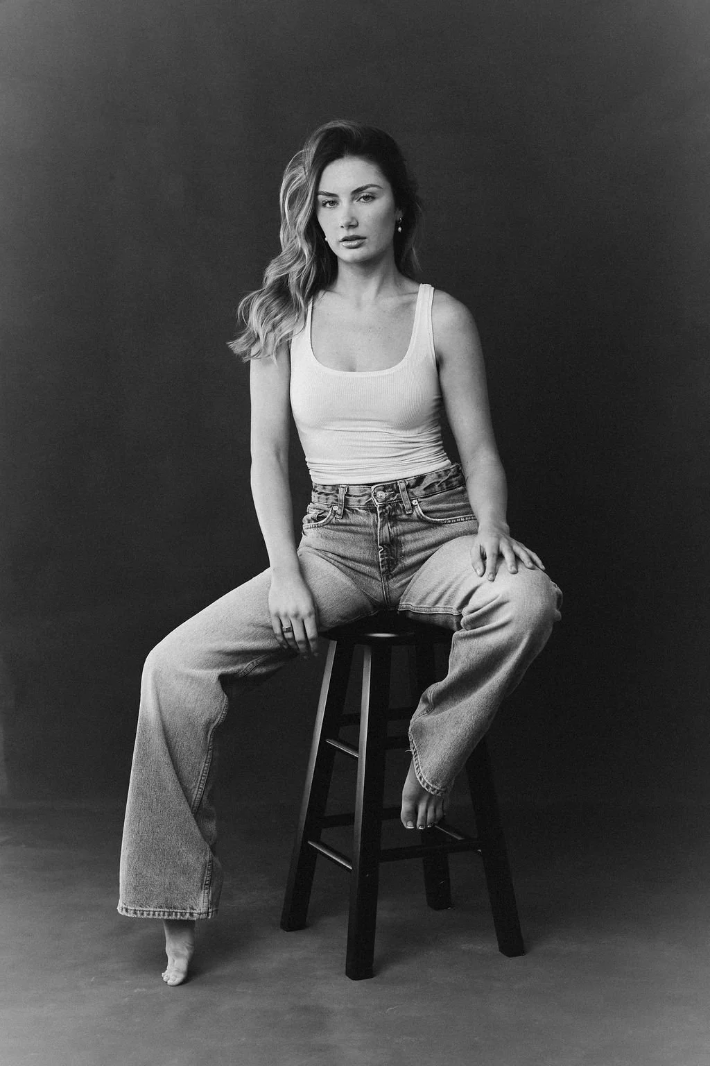 A young woman sitting on a stool with one foot on the ground and the other leg draped over the stool, against a dark background. She has wavy hair and is wearing a tank top and jeans.