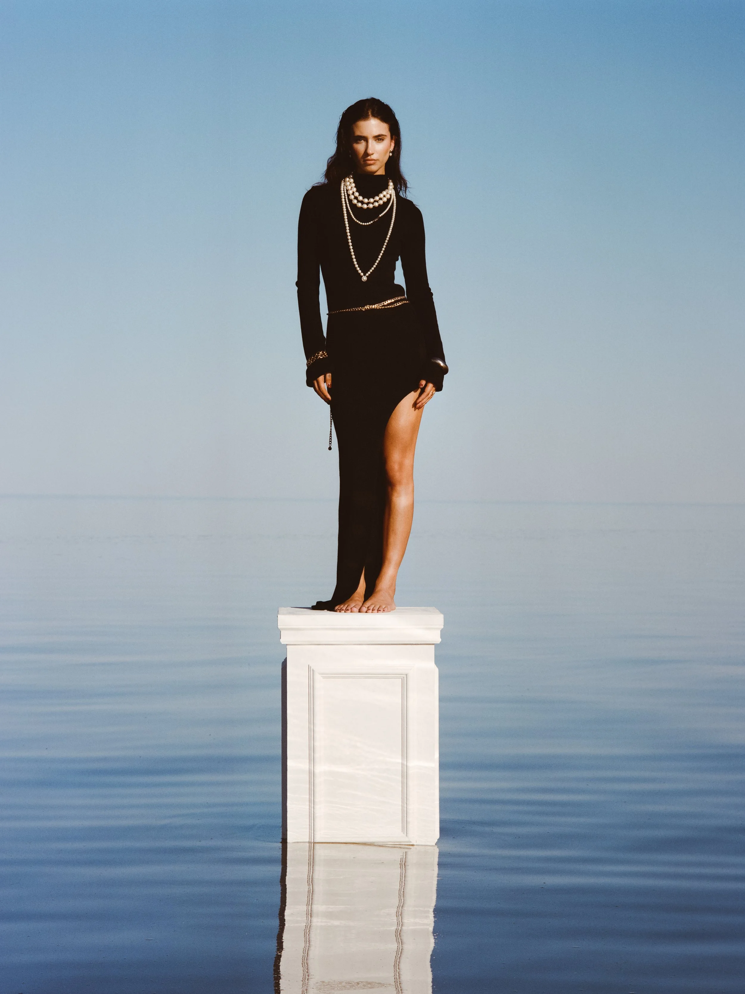 A woman dressed in black, adorned with pearl necklaces, standing barefoot on a white pedestal in a body of water with a clear blue sky background.