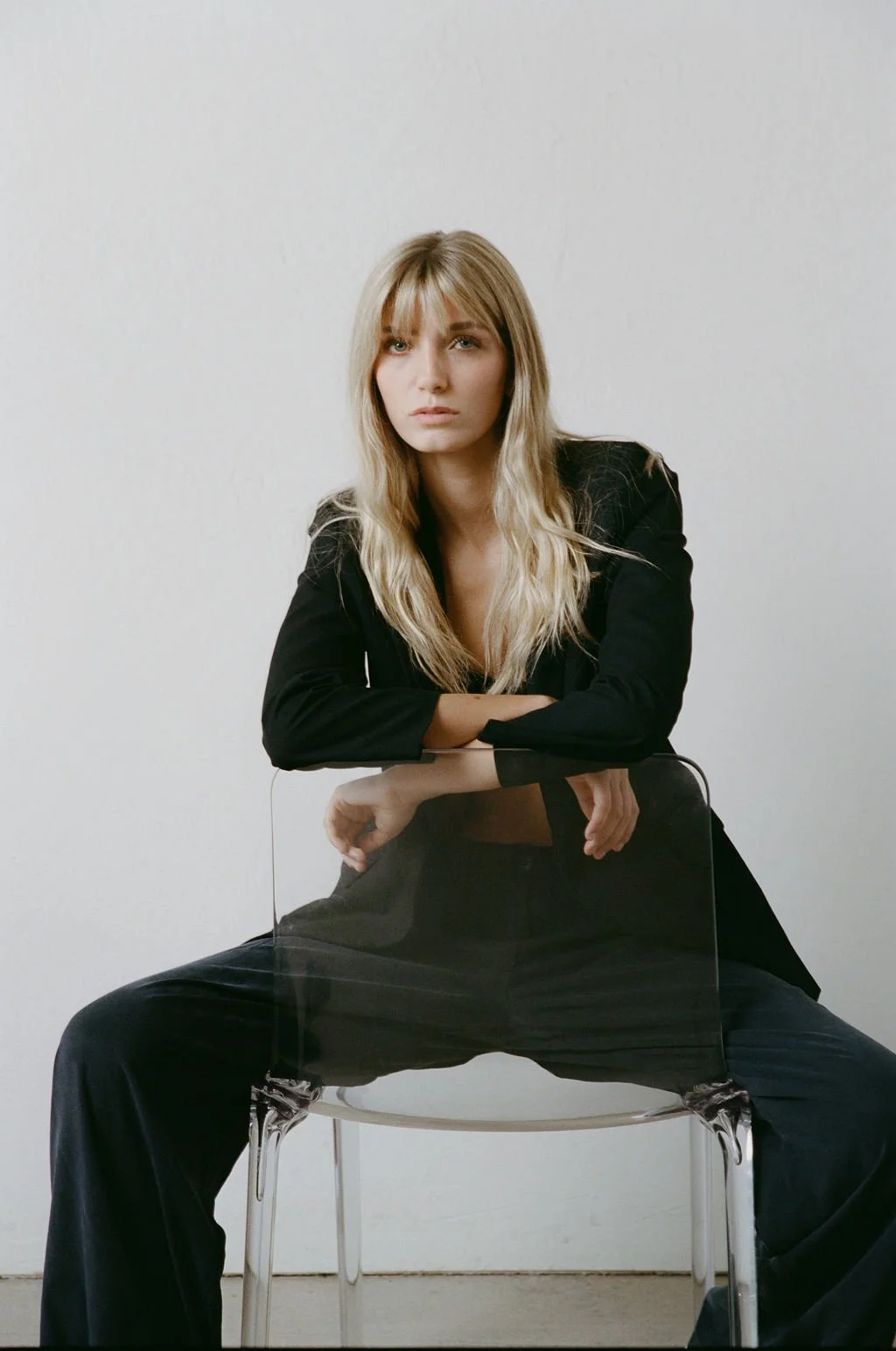 A woman with long blonde hair sitting on a clear chair, wearing a black blazer and black pants, posed with her legs apart and hands resting on her knees, against a plain white wall.