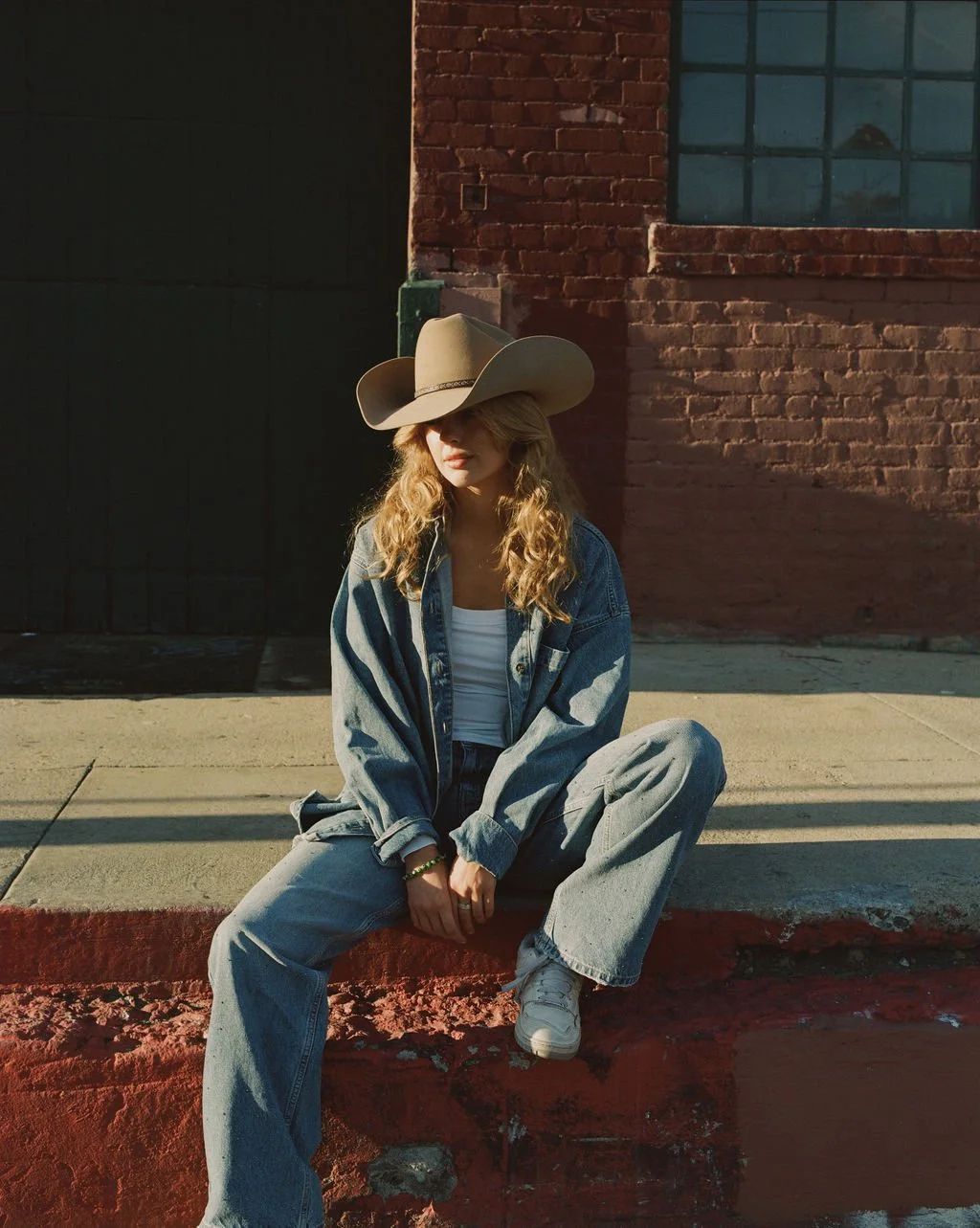 A young woman with curly blonde hair wearing a large cowboy hat, denim jacket, white top, and jeans, sitting on a red curb beside a brick building in the sunlight.