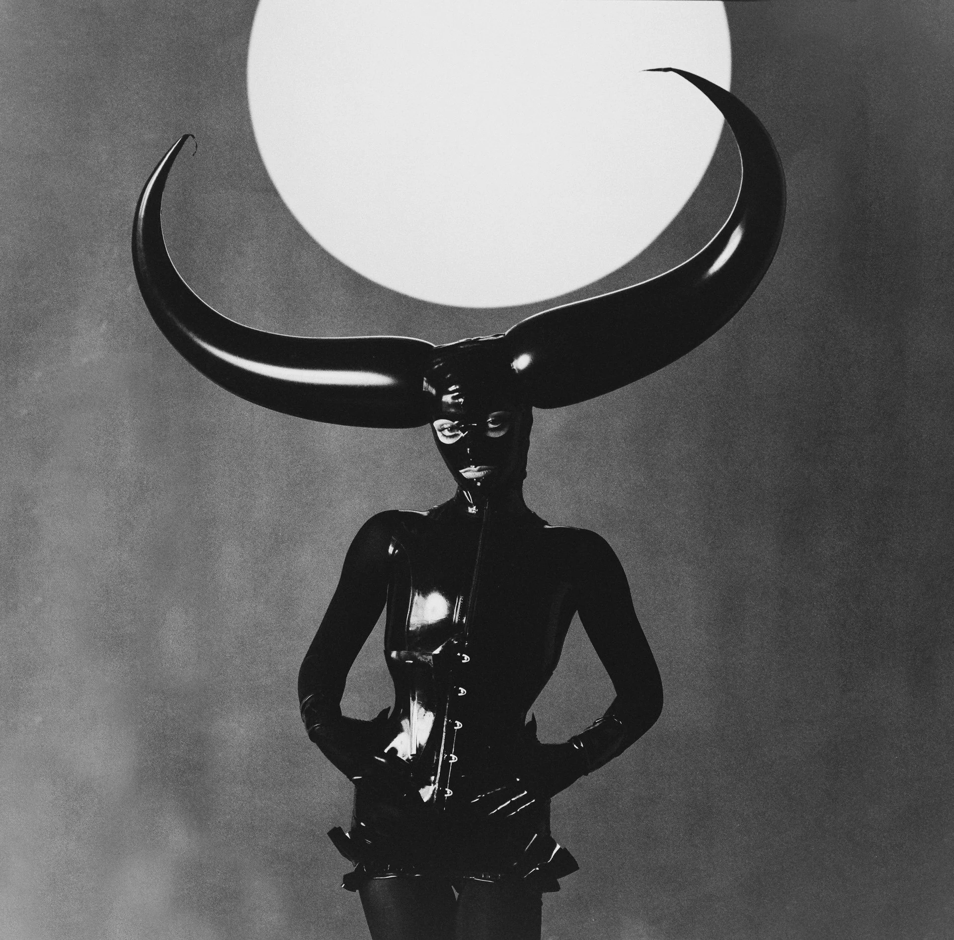 A person dressed in a shiny, black latex bodysuit with a large crescent moon headpiece under a bright full moon in a dark sky, all in black and white.