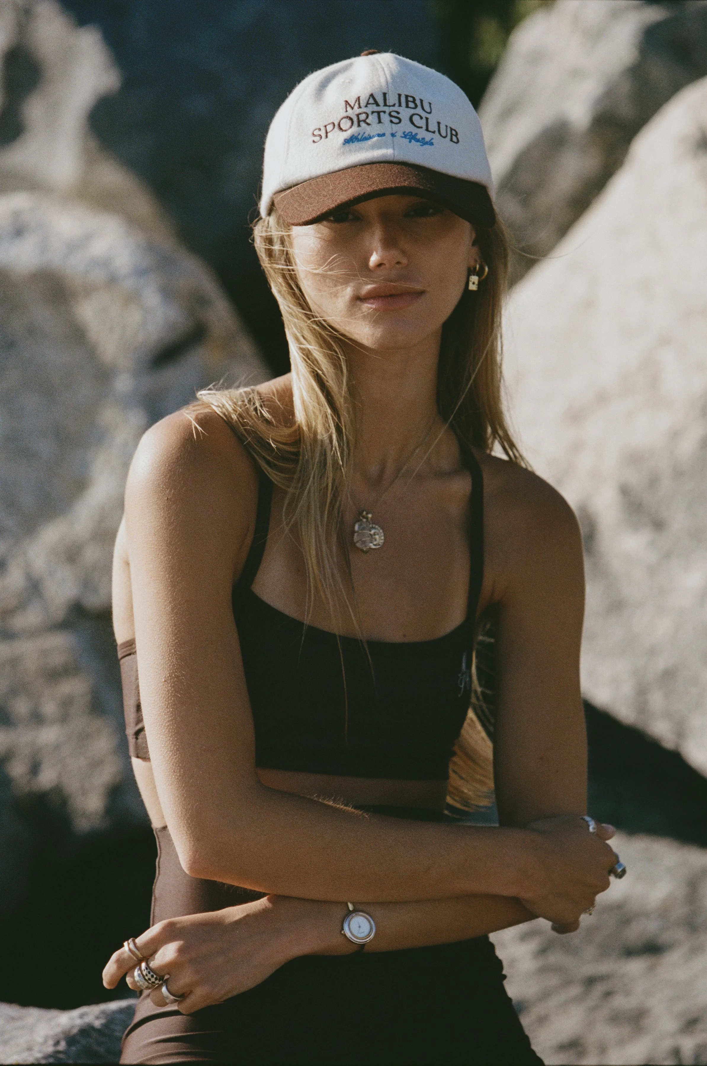 A woman wearing a Malibu Sports Club cap, a black sleeveless top, and jewelry, standing outdoors among rocks with sunlight illuminating her face.