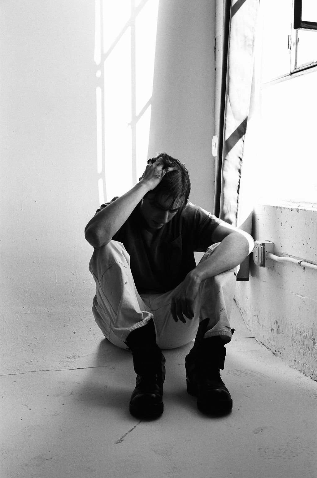 A young man with dark hair, wearing a dark t-shirt and light-colored pants, sitting on the floor with one hand on his head, in a room with natural light coming through a window.