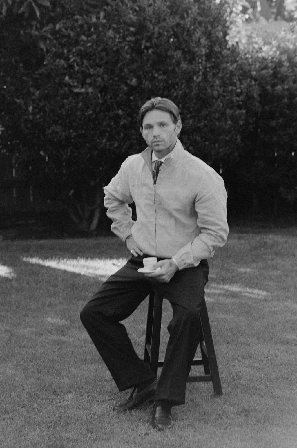 A man sitting on a stool outdoors, holding a cup, wearing a jacket and dress shoes, with trees in the background.