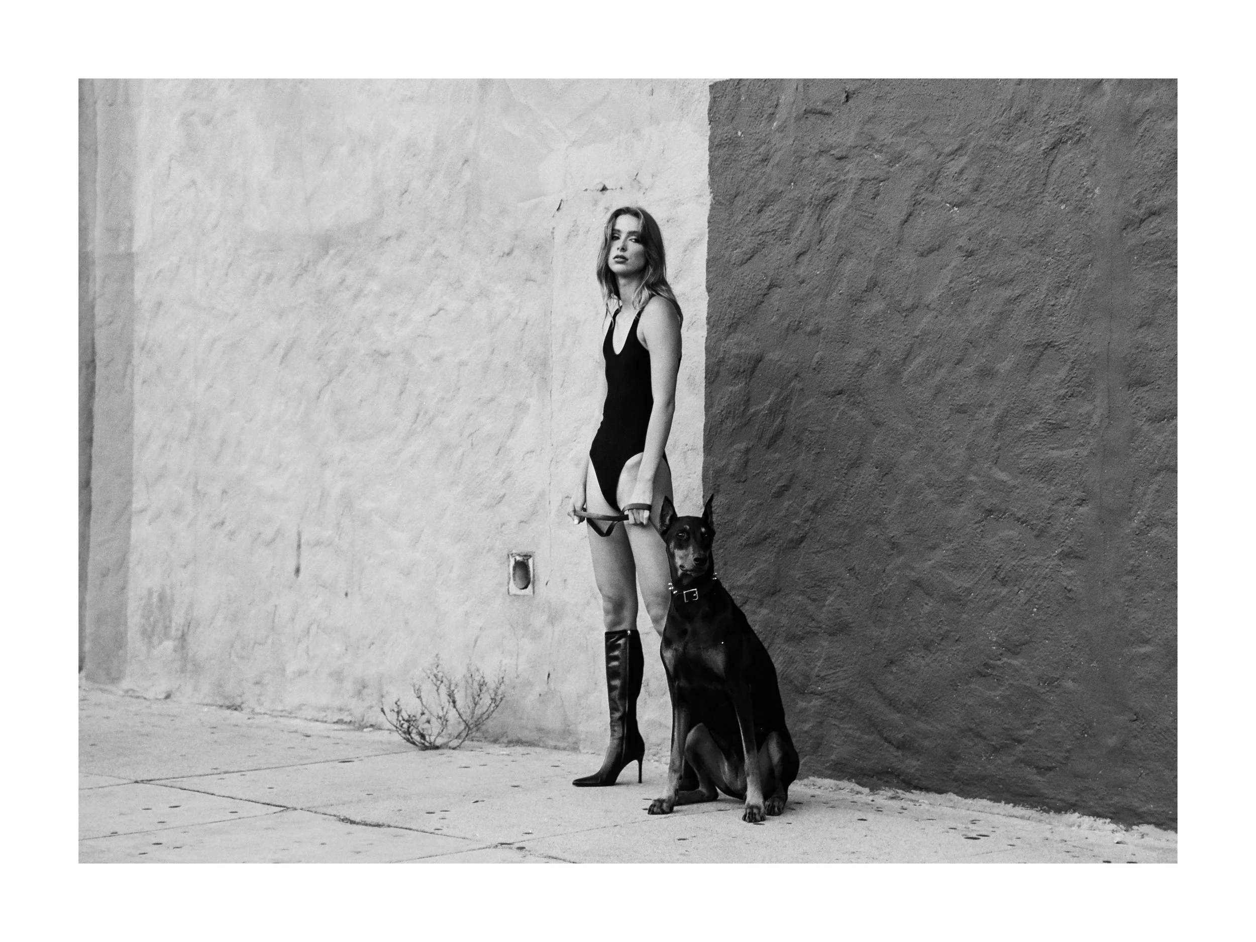 A woman in a black dress and high boots standing with a Doberman in front of a textured wall.