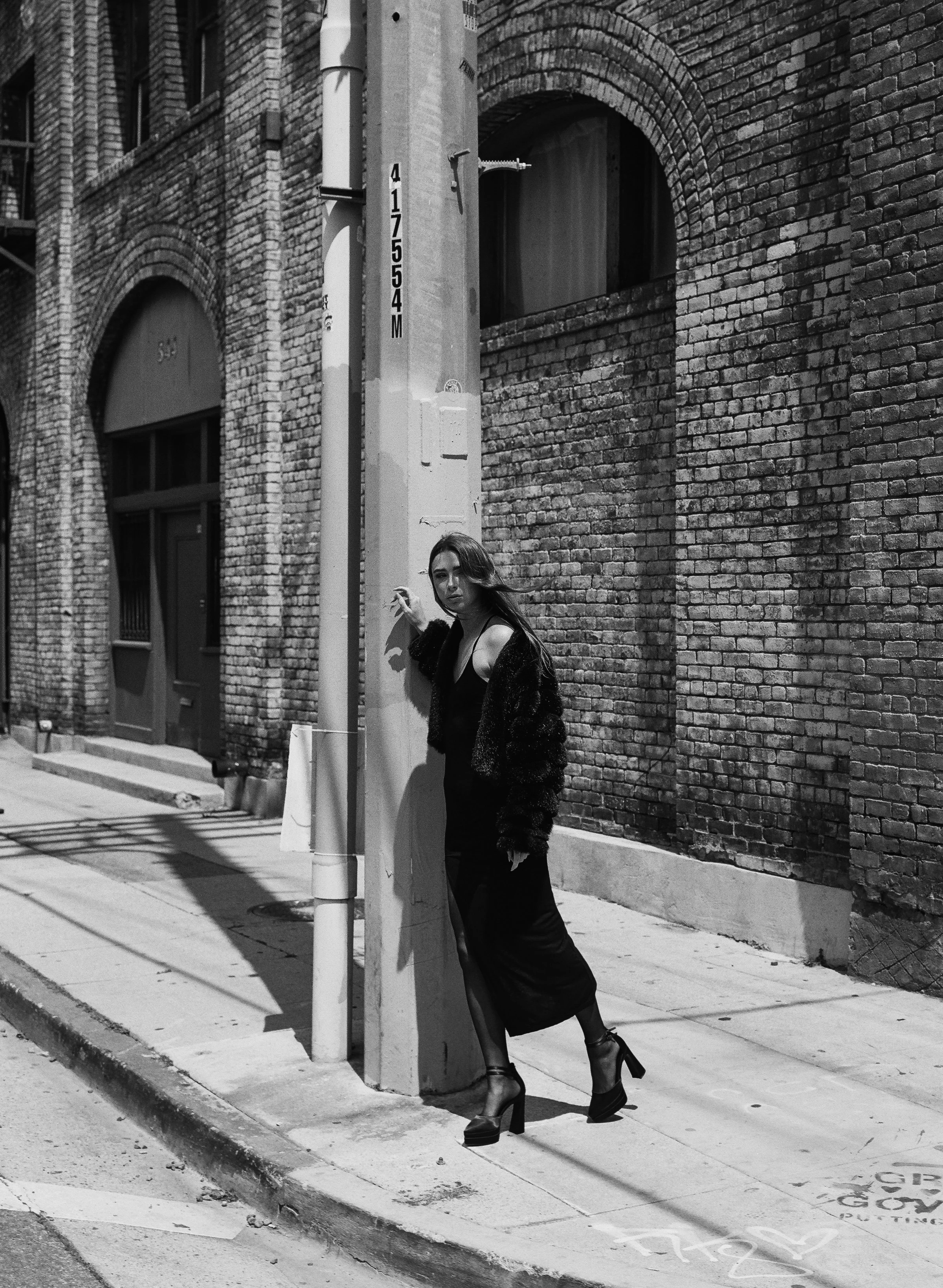 A woman in a long dress and high heels standing against a lamp post on a city sidewalk with brick buildings in the background, captured in black and white.