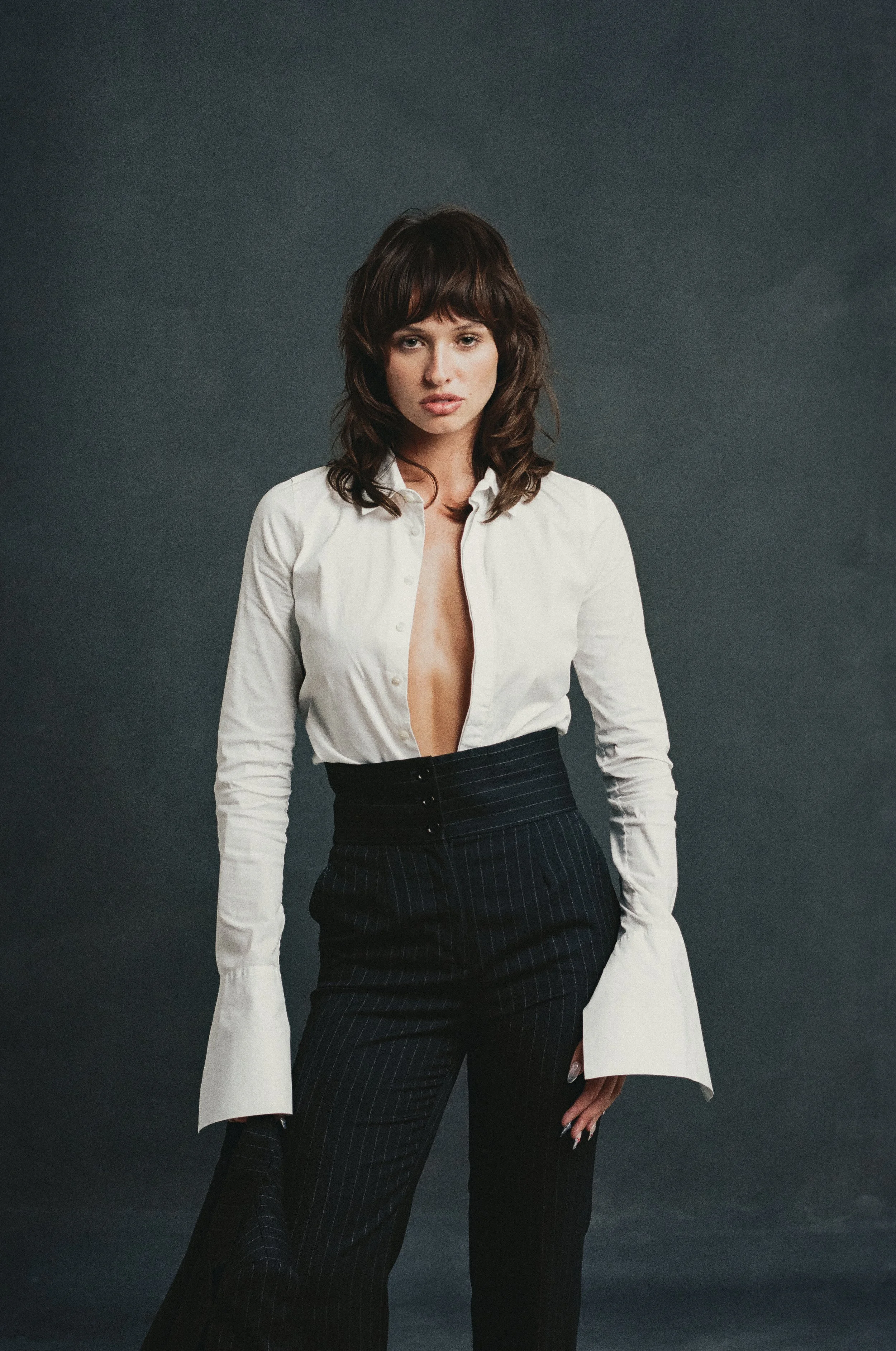 A woman with shoulder-length brown hair wearing a white button-up shirt with the top buttons undone and black high-waisted pinstripe pants standing against a dark background. Tatianna Ringsby