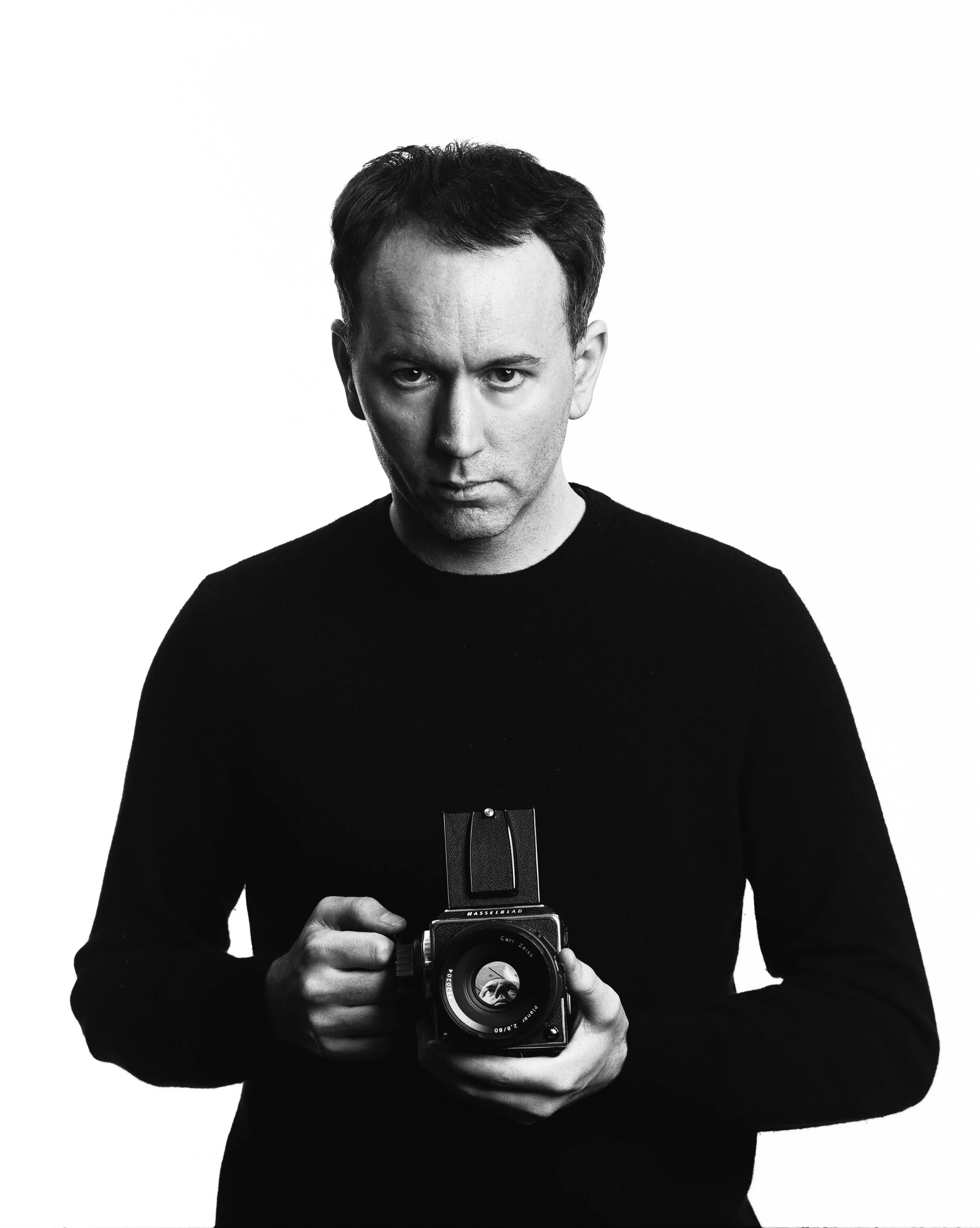A man with dark hair and wearing a black shirt looking directly at the camera, holding a vintage camera with a flash attachment, black and white photo.