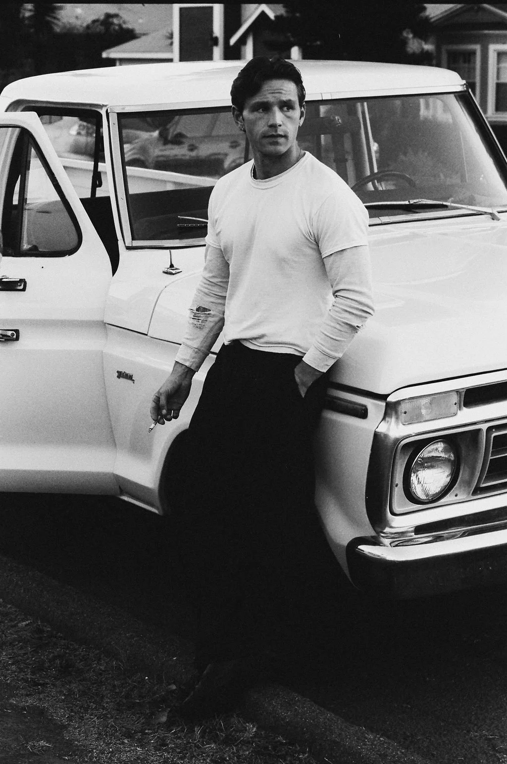 A man wearing a white long-sleeve shirt and dark pants standing next to a vintage truck, holding a cigarette in his right hand and looking to his left, in a residential neighborhood.