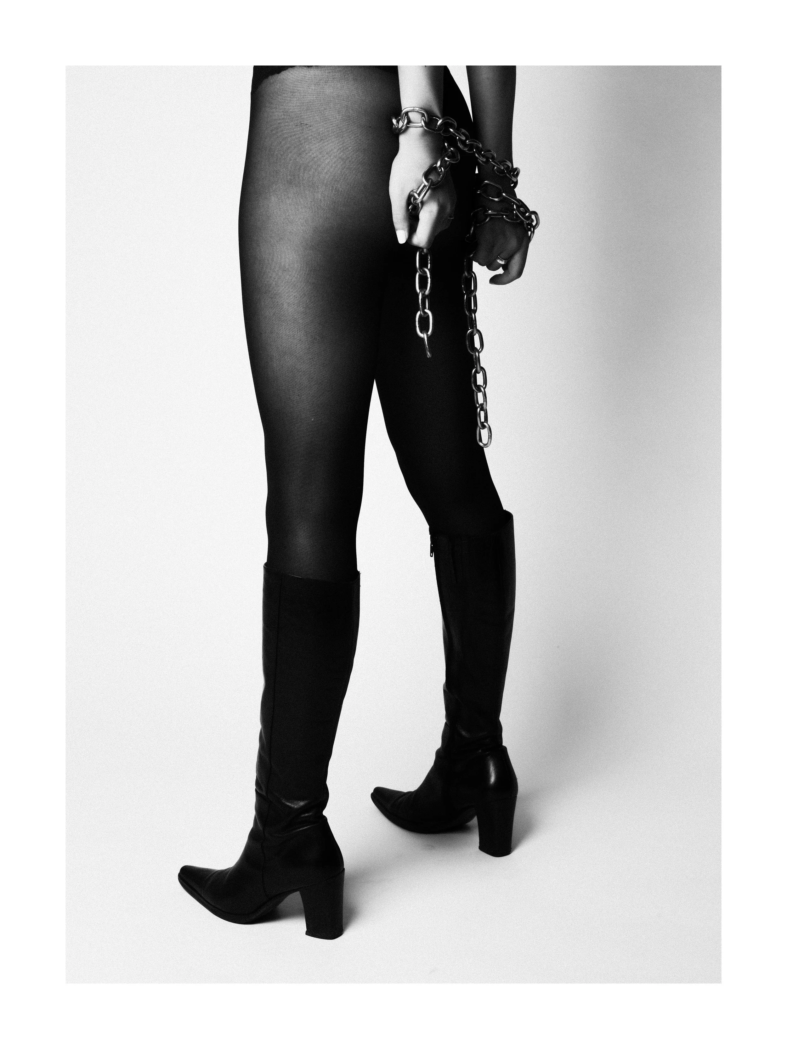 A person wearing black thigh-high boots, black sheer tights, and black gloves, with hands restrained by a chain.