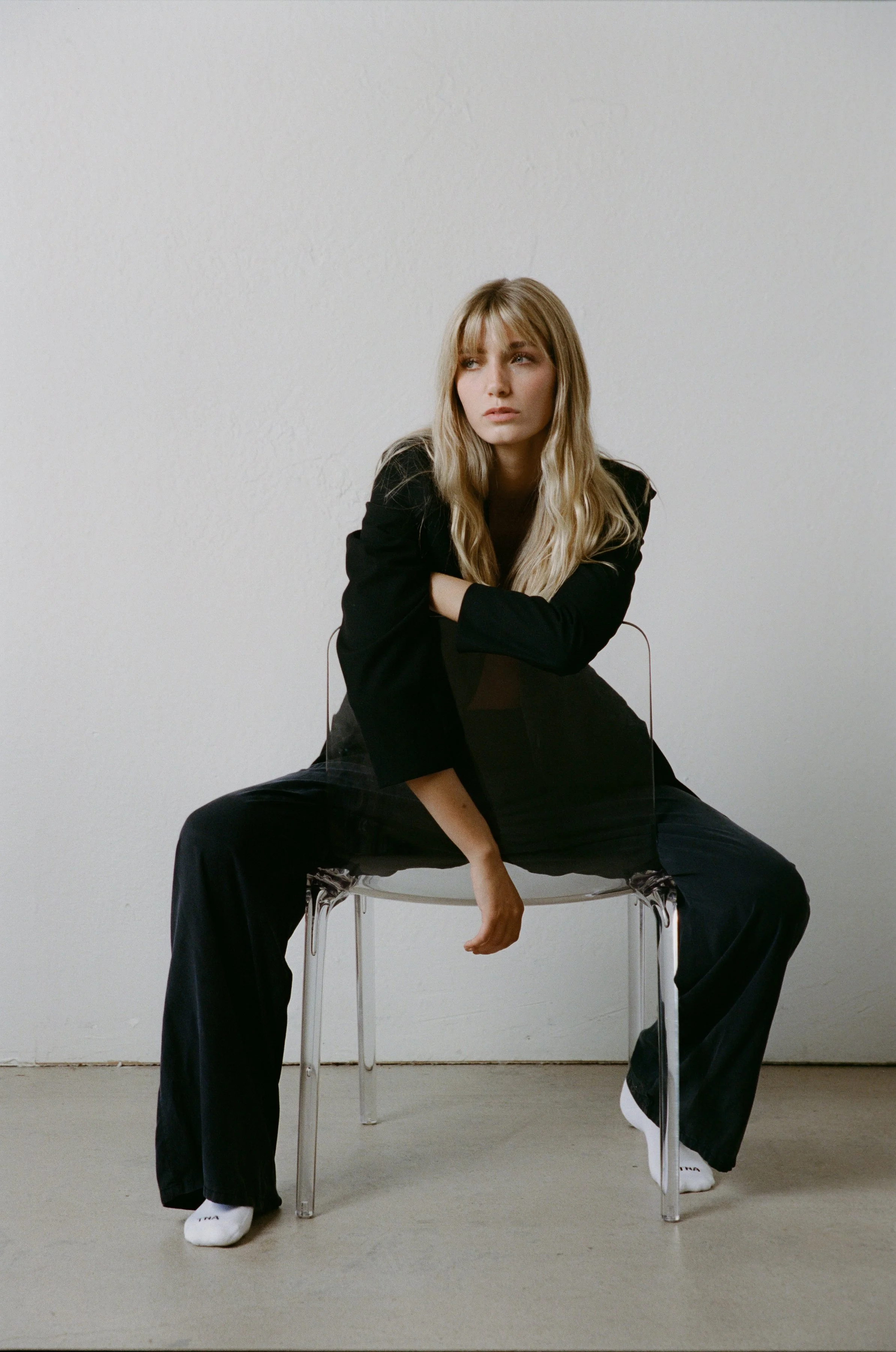 A woman with long blonde hair wearing a black blazer and black wide-leg pants sitting on a clear acrylic chair against a plain white wall, looking thoughtfully to the side.