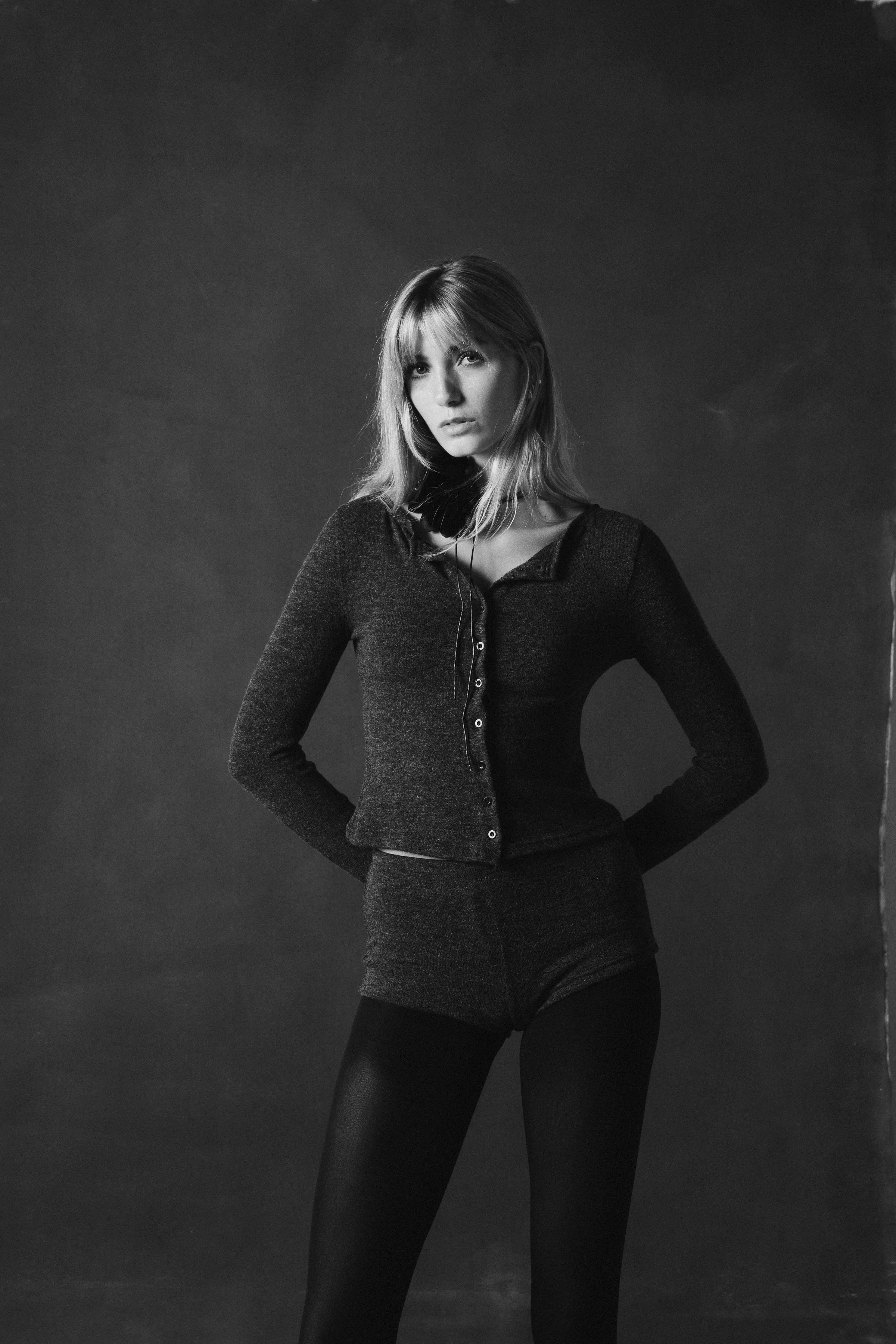 A young woman with blonde hair wearing a dark long-sleeve top, shorts, and tights in a black-and-white studio portrait.