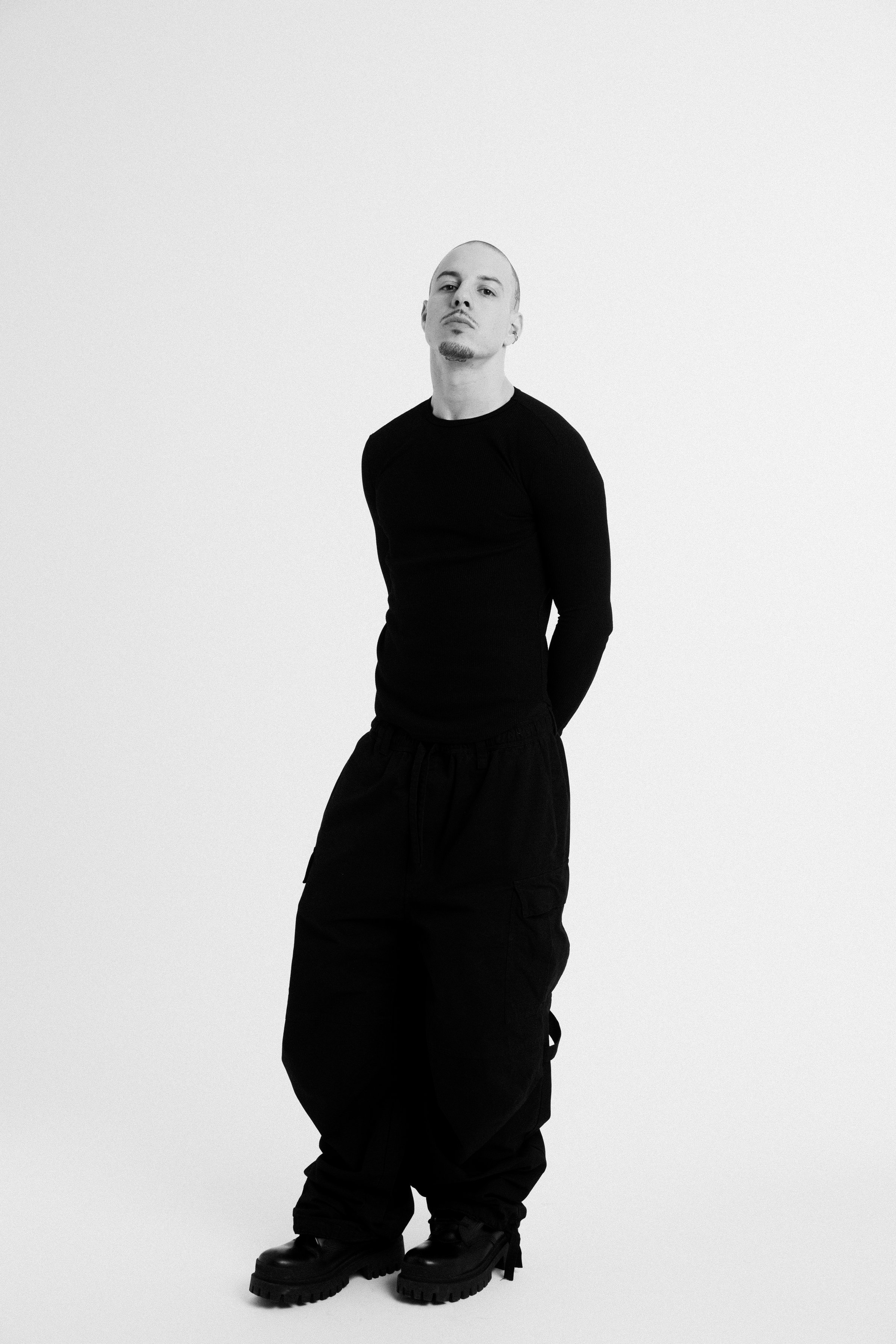 A man standing against a plain light background, dressed in black, with a serious expression, black pants, and black shoes.