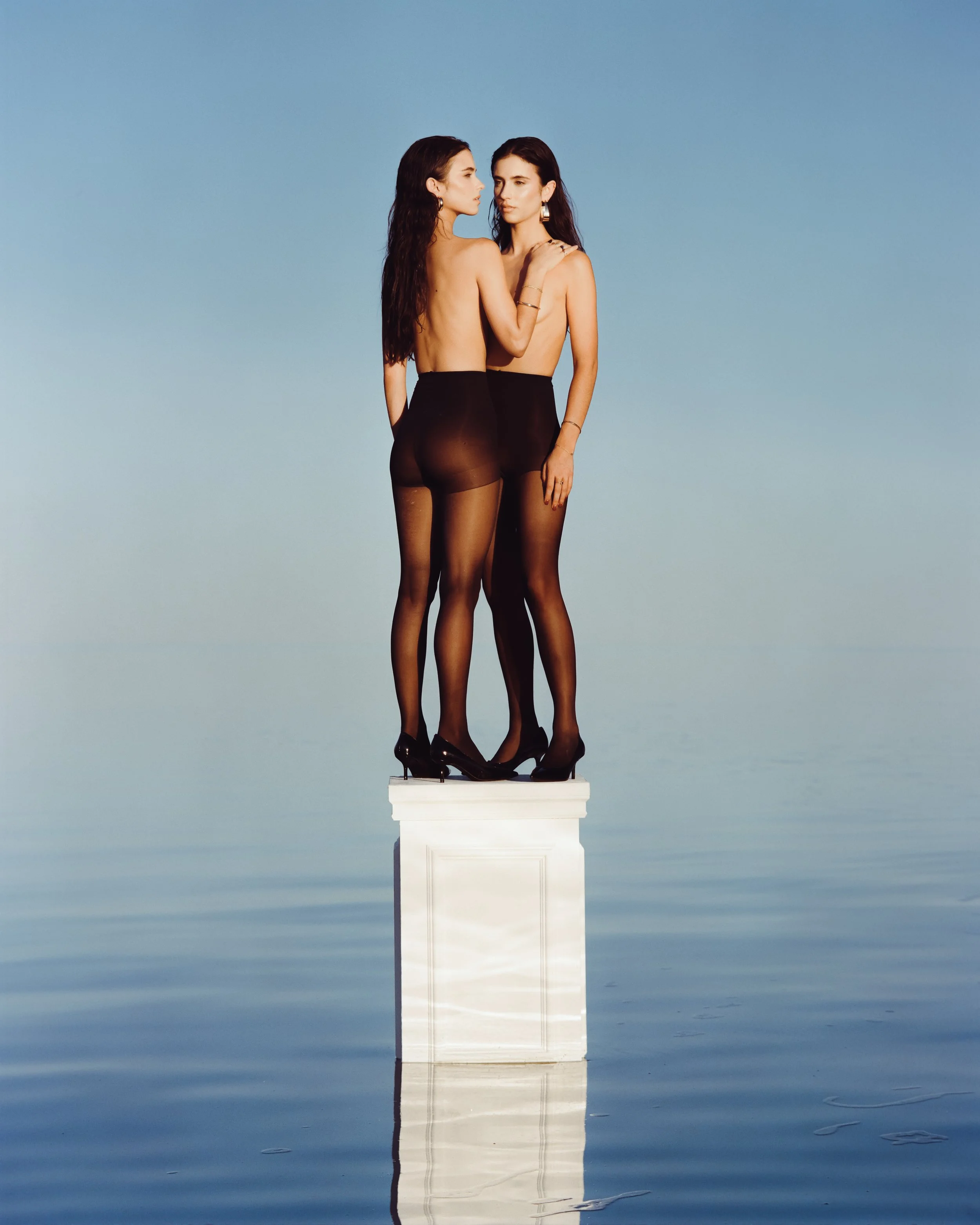 Two women in black stockings and black high heels standing on a white pedestal in water with a clear blue sky background.