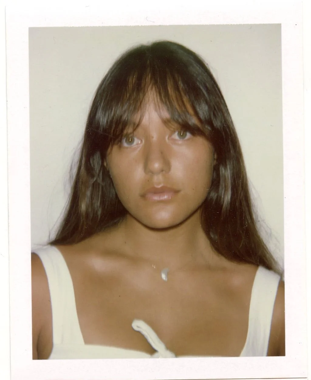 A woman with long brown hair and bangs wearing a white tank top and a small necklace, looking directly at the camera against a plain background.