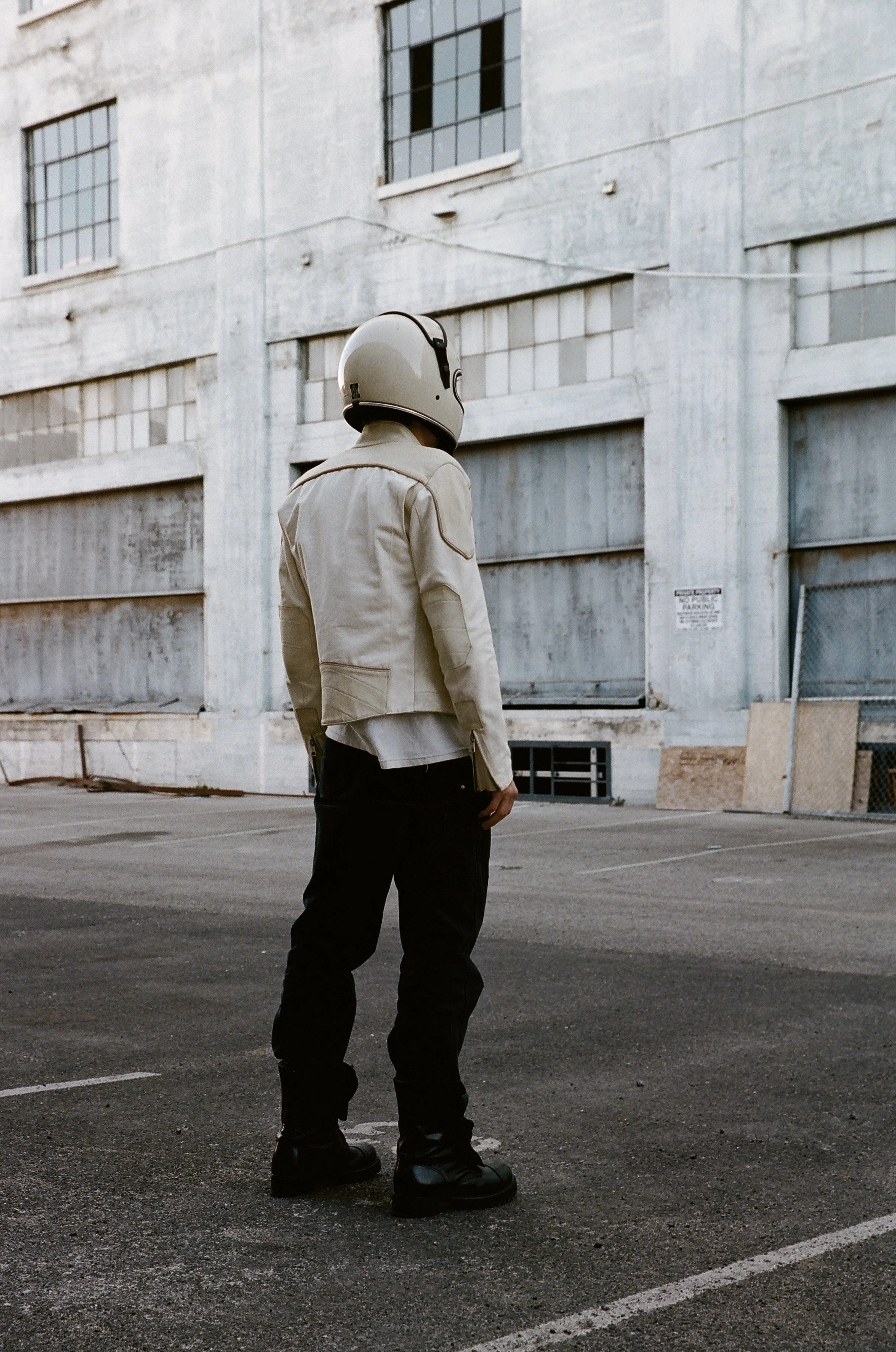 Person wearing a helmet and beige jacket standing face down in an empty parking lot, an abandoned building in background.