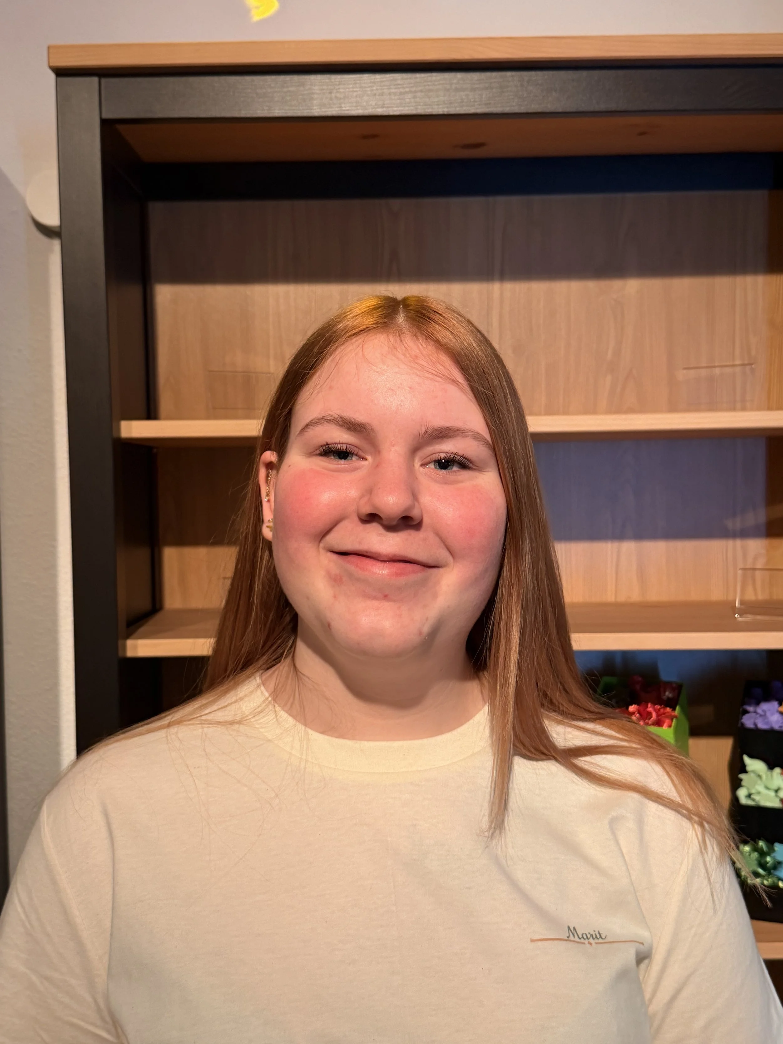 Ein lächelndes Mädchen mit braunen Haaren, das vor einem Holzbücherregal steht.