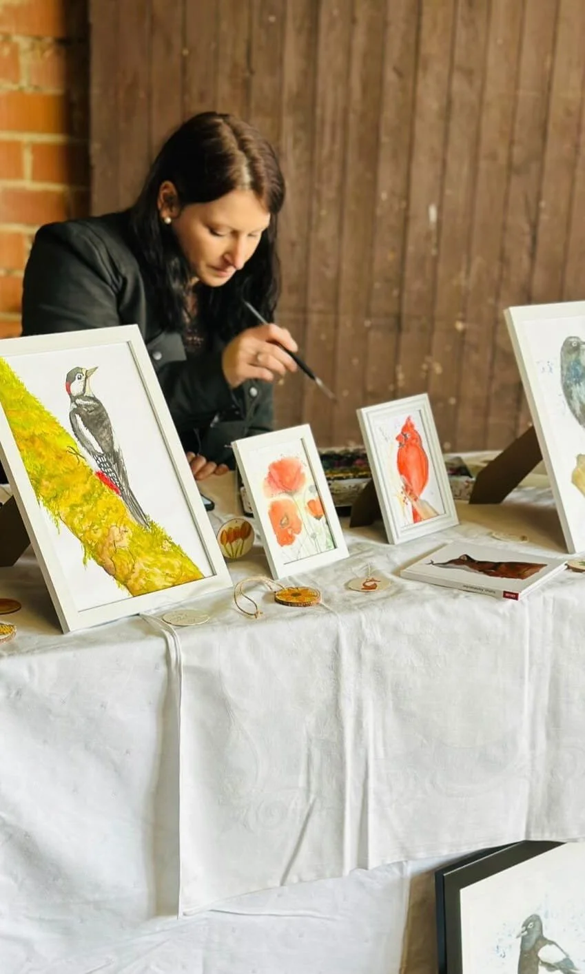 Frau beim Malen von Wasserbildern mit Vogelmotiven an einem Tisch mit verschiedenen Kunstwerken und Dekorationen.