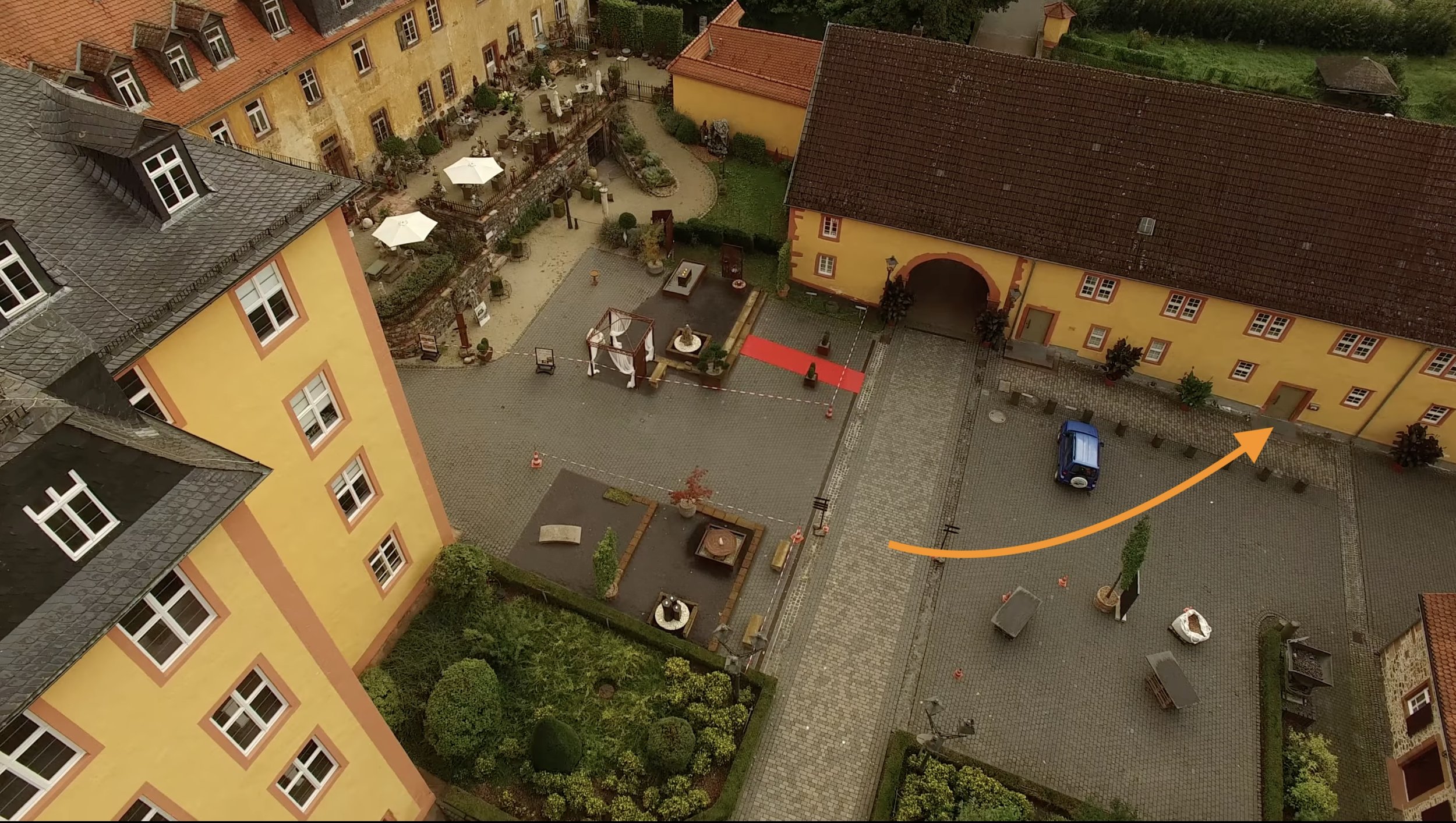 Ein Blick von oben auf einen historischen Hof mit gelben Gebäuden, einem gepflasterten Platz, einigen Bäumen und einer kleinen Gartenfläche mit Sitzgelegenheiten. Es gibt auch eine kleine blaue Fahrzeuge und Absperrbänder, die einen Bereich markieren.