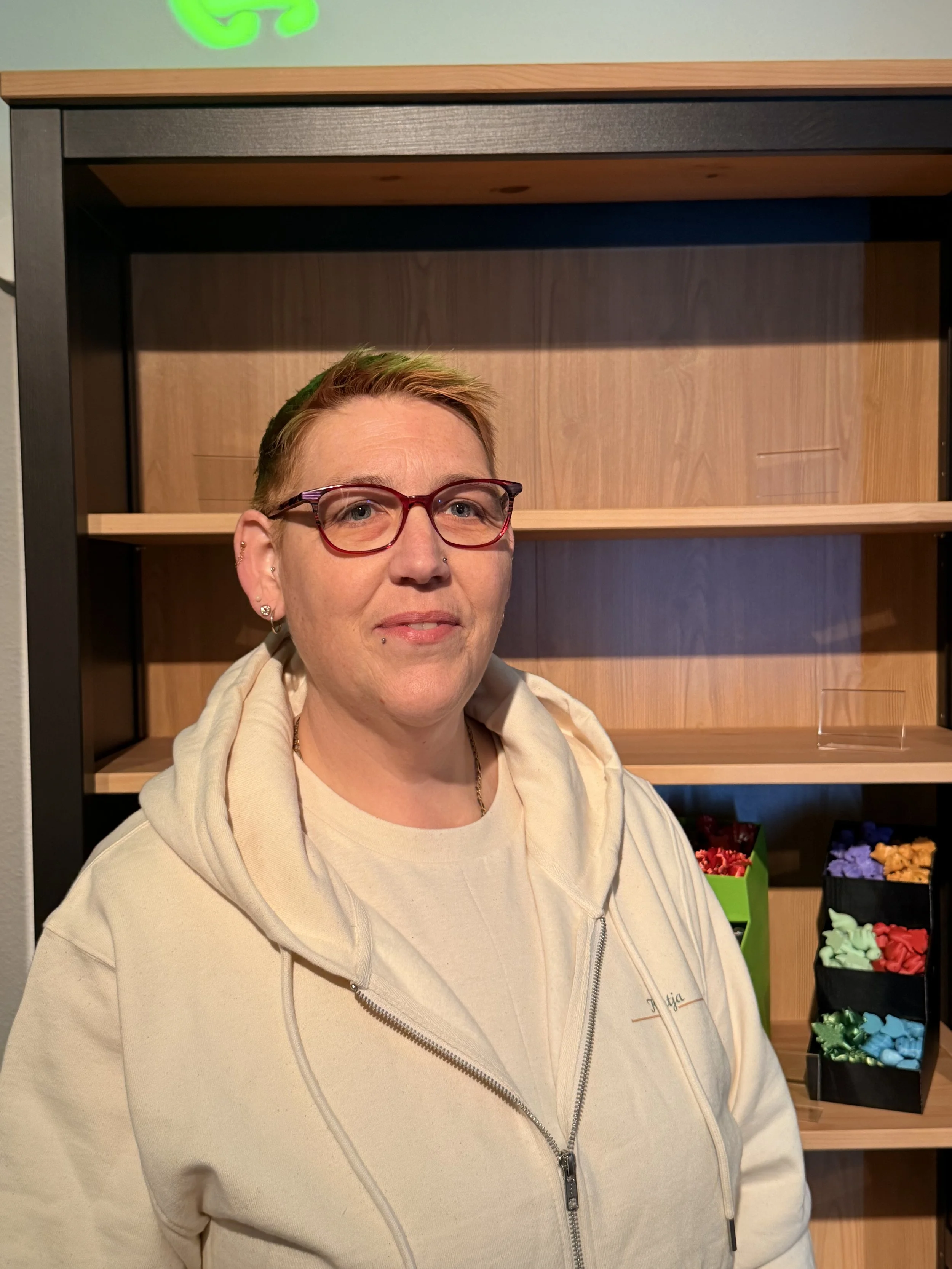 Eine Person mit kurzen braunen Haaren, Brille und einem hellen Hoodie steht vor leeren Holzregalen.