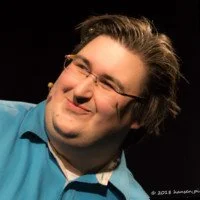 Ein Mann mit Brille und dunklen Haaren, lächelnd, trägt ein blaues Hemd, vor dunklem Hintergrund.