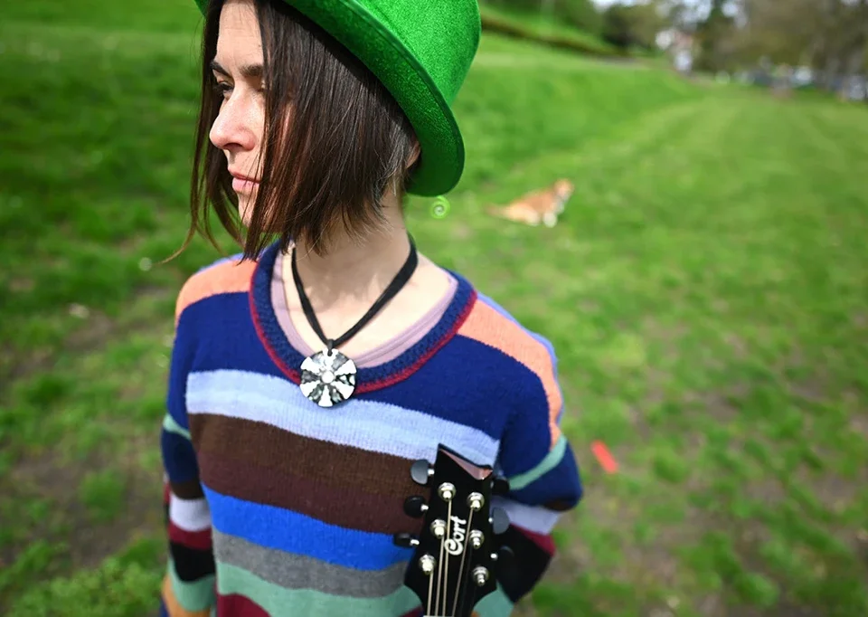 Junge Frau (Pauli) trägt grünen Hut und bunte gestreifte Pullover, hält eine Gitarre, im Hintergrund ein Hund auf grünem Rasen im Freien.