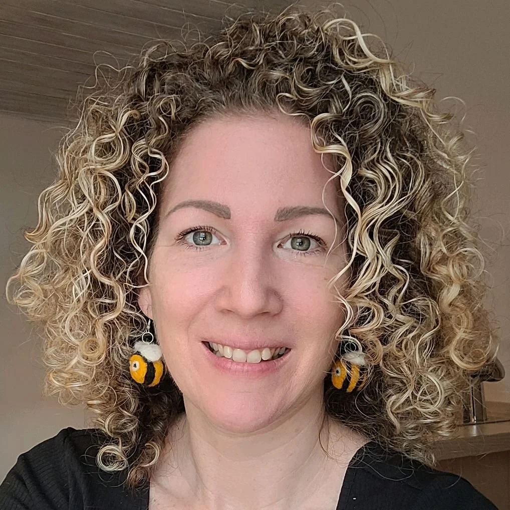 Nahaufnahme einer lachenden Frau mit lockigem, blondem Haar, blauen Augen und Bienen-Ohrringen.