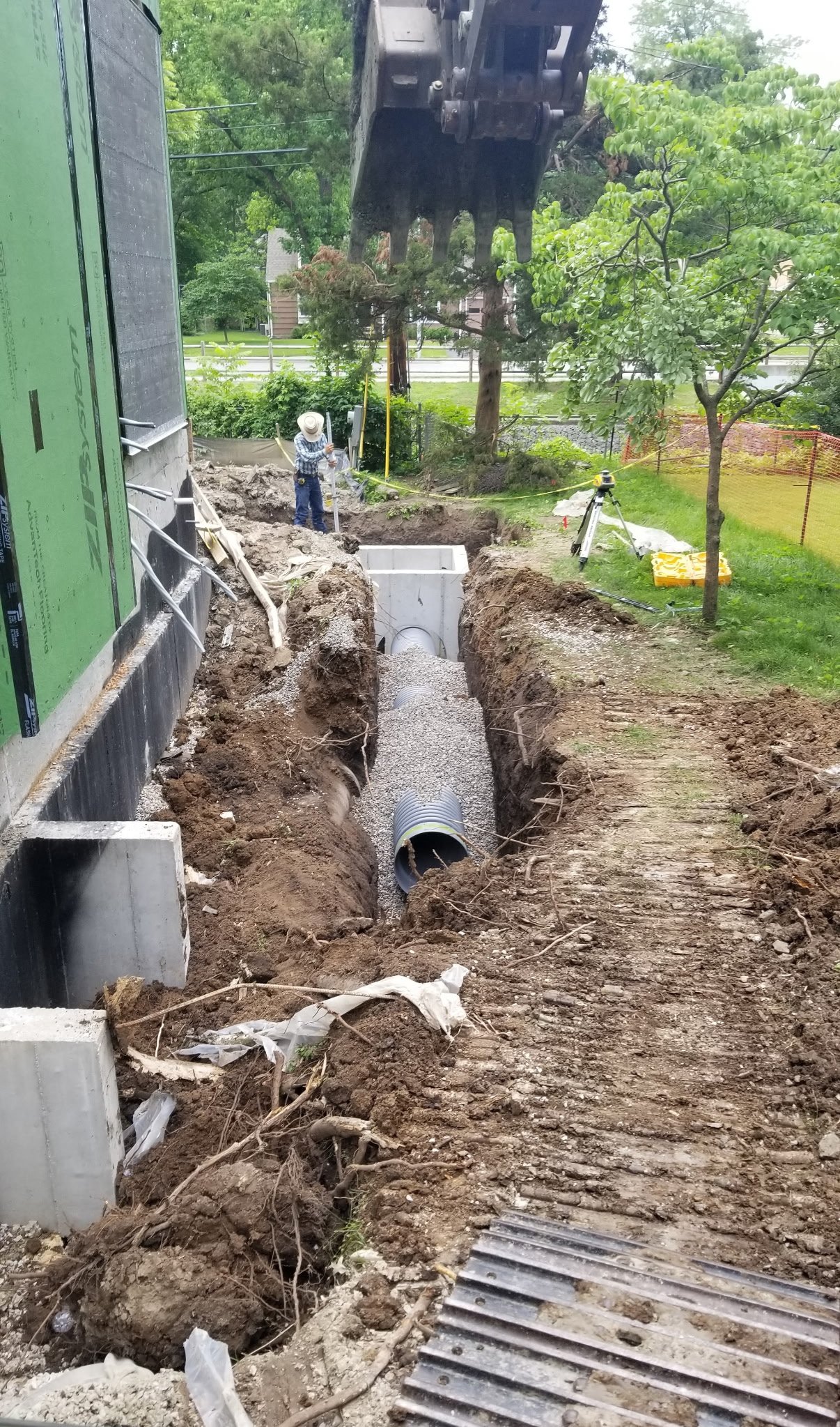 Construction workers install underground piping in a trench outside a building, with construction equipment and tools visible.
