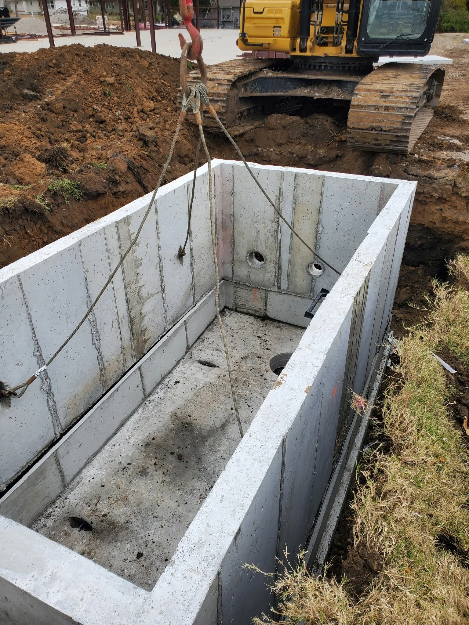 A concrete foundation with drainage pipes inside a construction site, with a yellow excavator and dirt surrounding it.