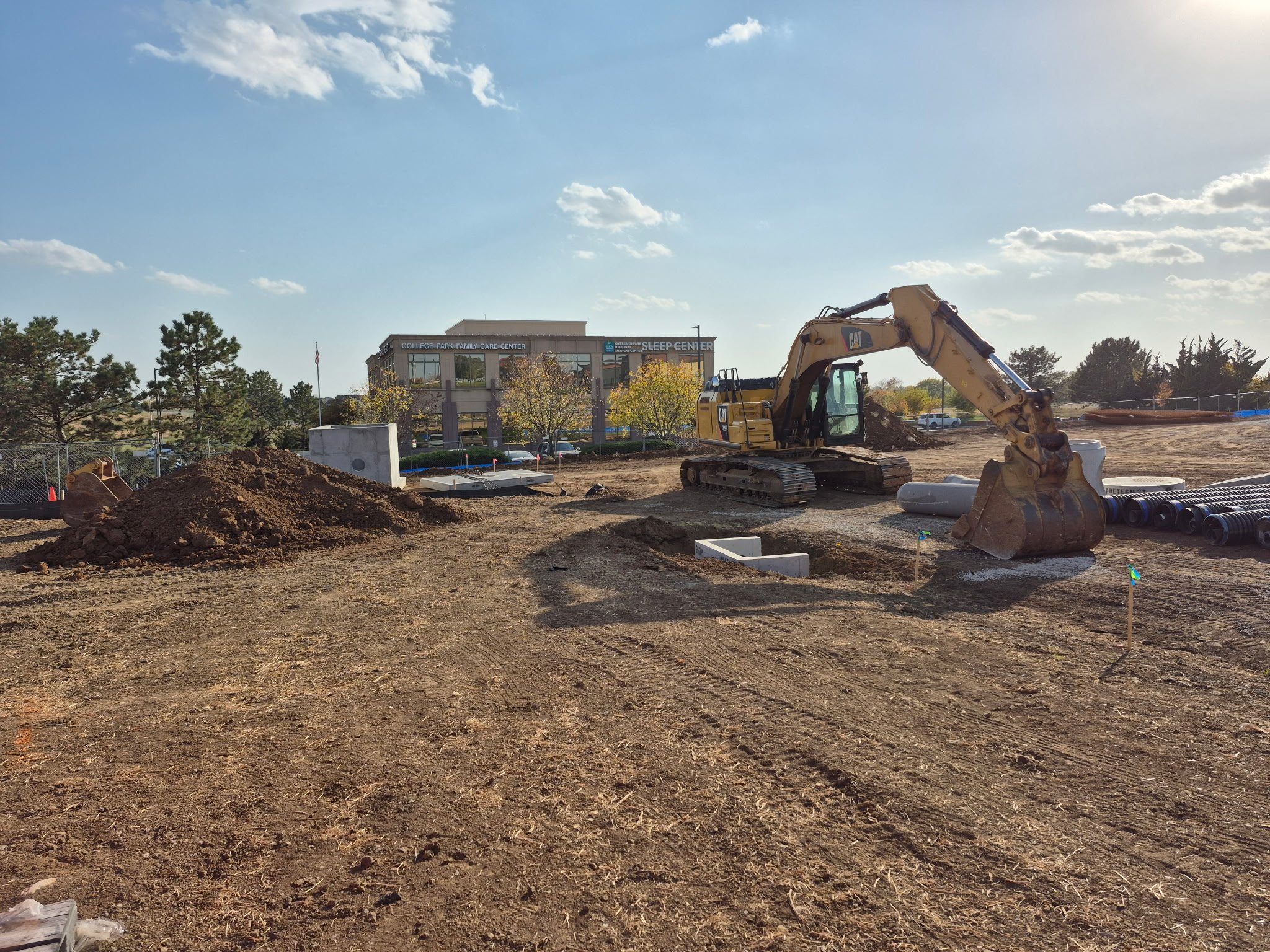 Construction site with a yellow excavator moving dirt, piles of soil, and pipes on cleared land with a modern building and trees in the background under a partly cloudy sky.