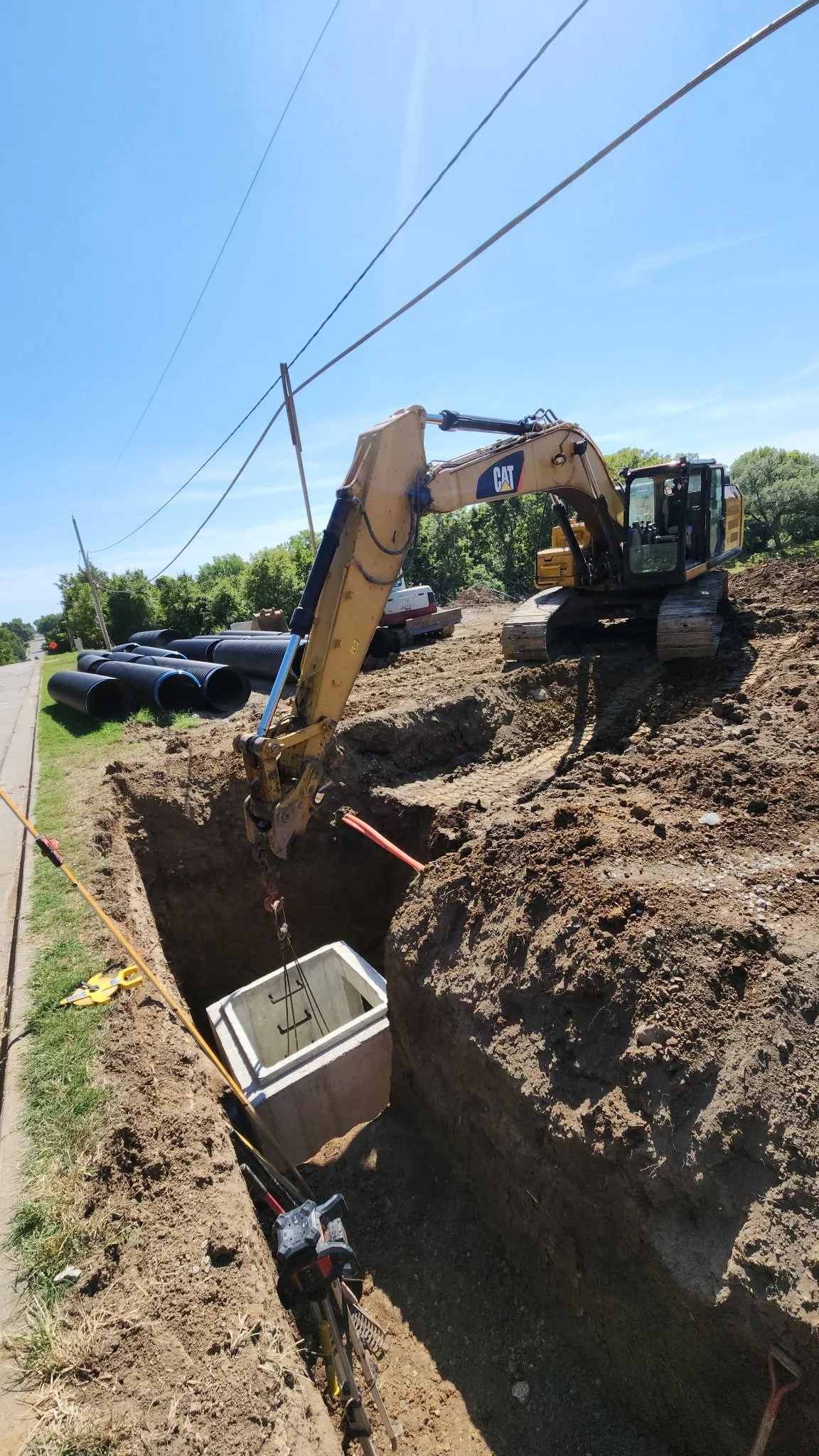 Construction site with a yellow Caterpillar excavator digging a trench for underground pipes next to a road, with large black pipes lying nearby under a clear blue sky.