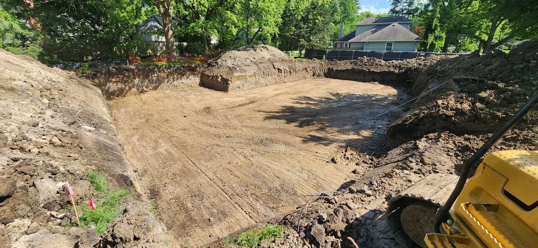 Construction site with excavated land, tire tracks, and a small yellow construction machine, surrounded by trees and neighboring houses.