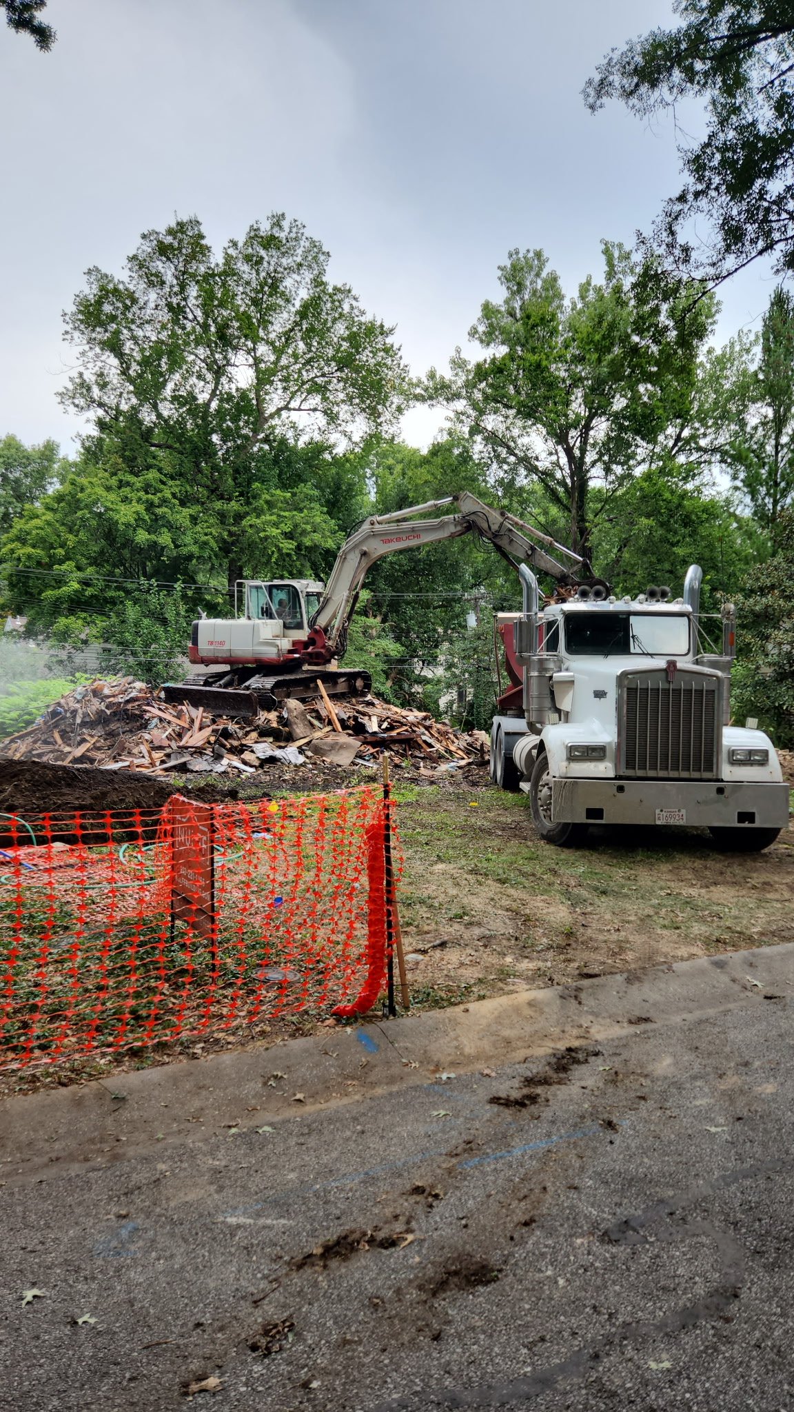 Construction site with a white dump truck and a small excavator working on a pile of dirt and wood debris, surrounded by green trees and an orange safety fence.